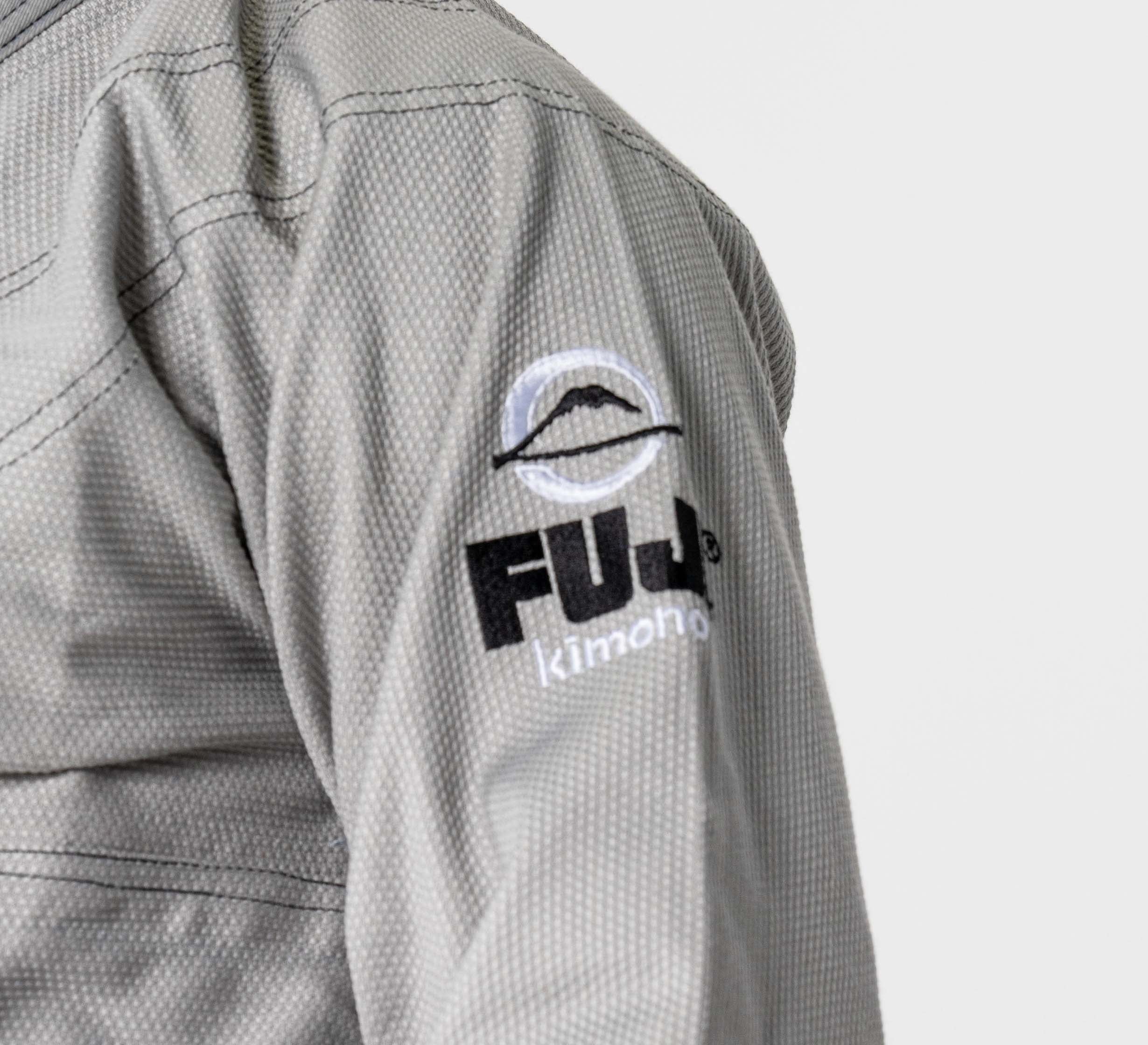Close-up of a person wearing the Lightweight BJJ Gi Grey, featuring a textured light gray fabric with the FUJI Kimono logo and a mountain graphic on the sleeve. The background is plain white.