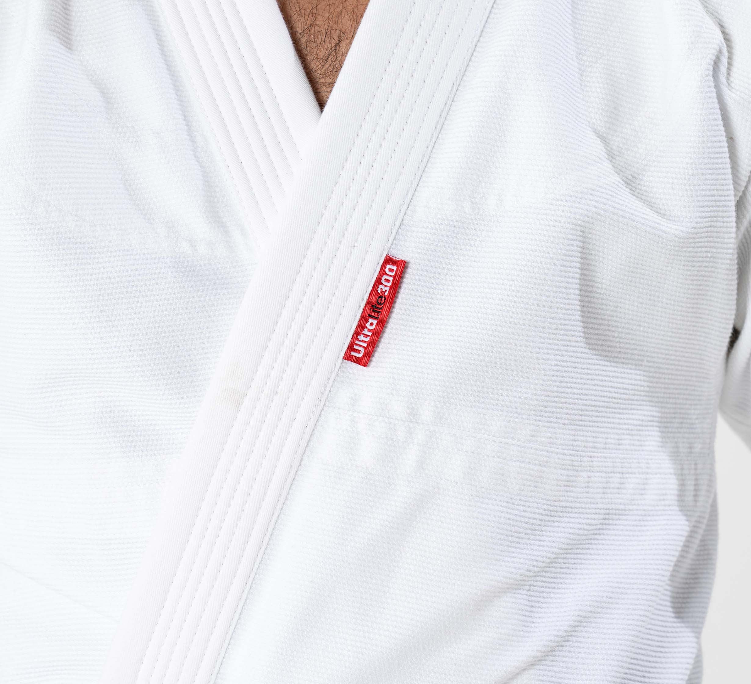 Close-up of a person wearing the Kids Ultra Lite 300 BJJ Gi in white, featuring a visible red Ultra Light 3.0 tag on the chest. The neck and part of the chest are also in view.