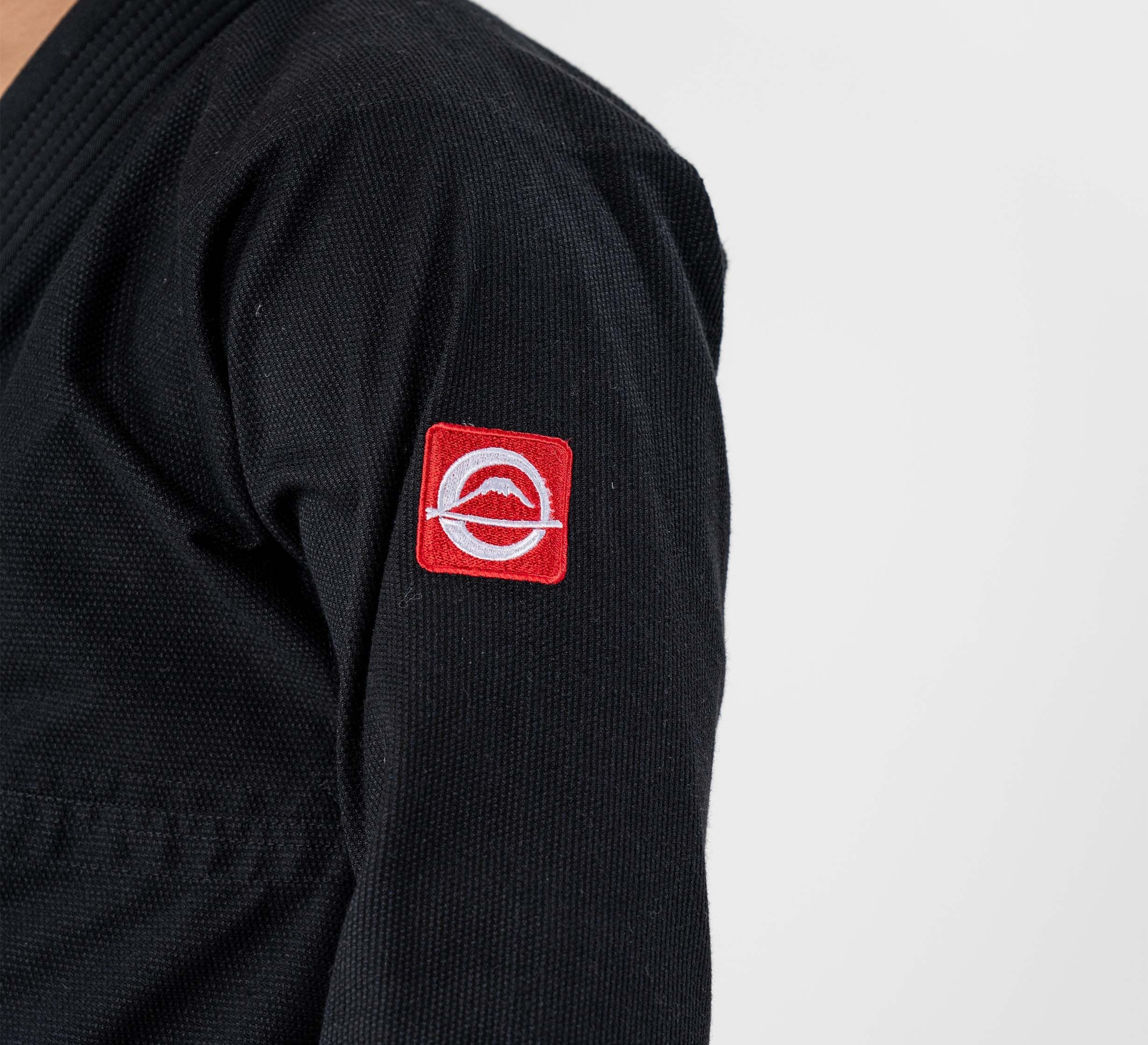 A person wears the Ultra Lite 300 BJJ Gi Black, featuring a red and white circular logo patch on the upper arm, against a plain white background.