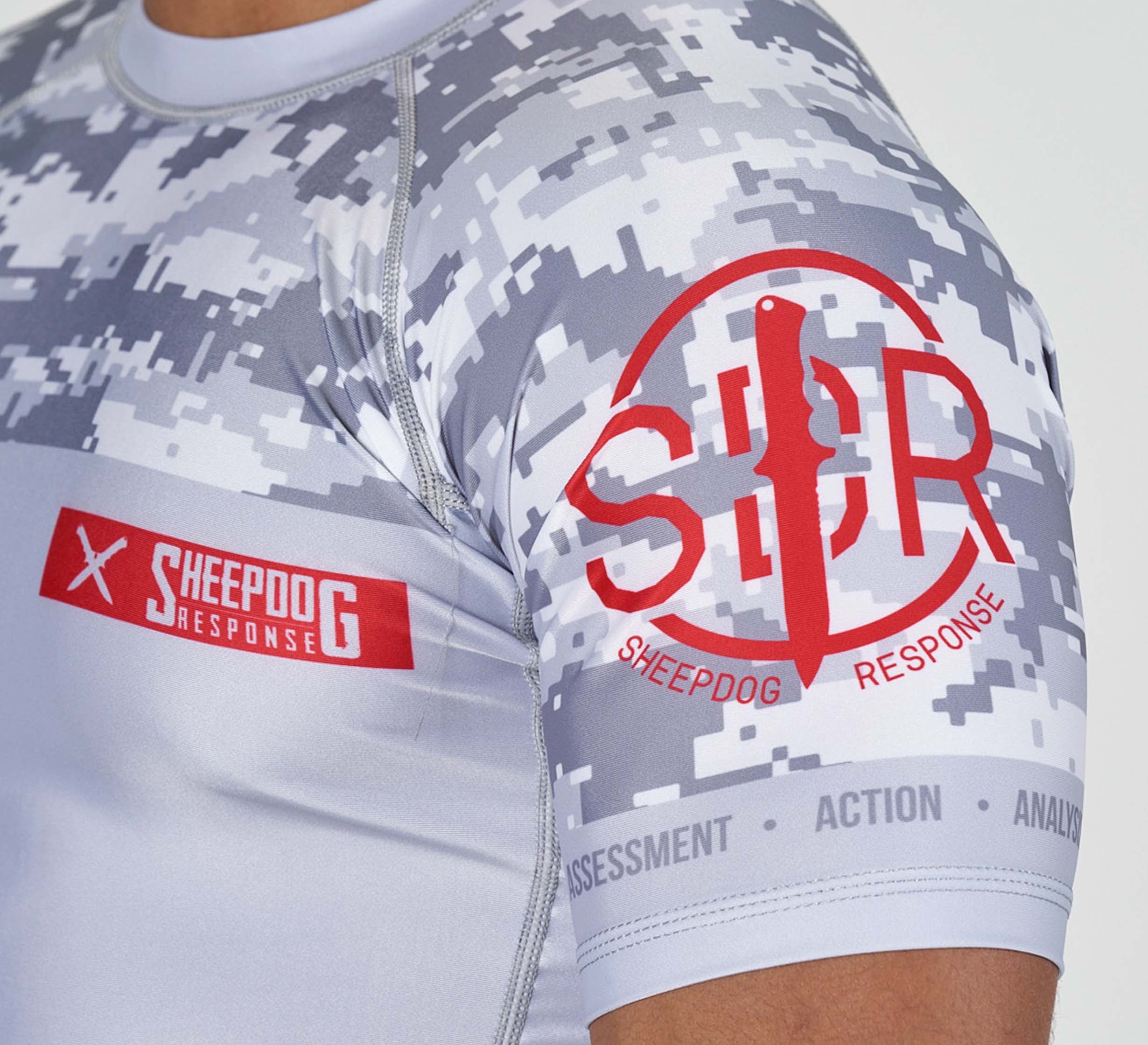 Close-up of a person wearing the Sheepdog Response Flex Lite Rashguard Grey, featuring a white and gray digital camo pattern with red Sheepdog Response logos and "Assessment Action Analysis" on the sleeve cuff.