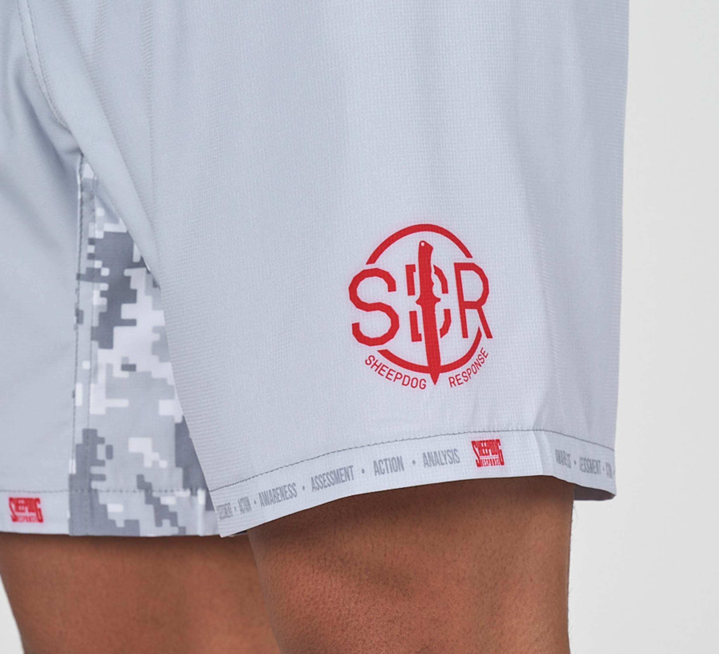 A close-up of a person wearing Sheepdog Response Flex Lite Shorts Grey, featuring a light gray hue, camouflage side panels, and a red Sheepdog Response logo near the hem.