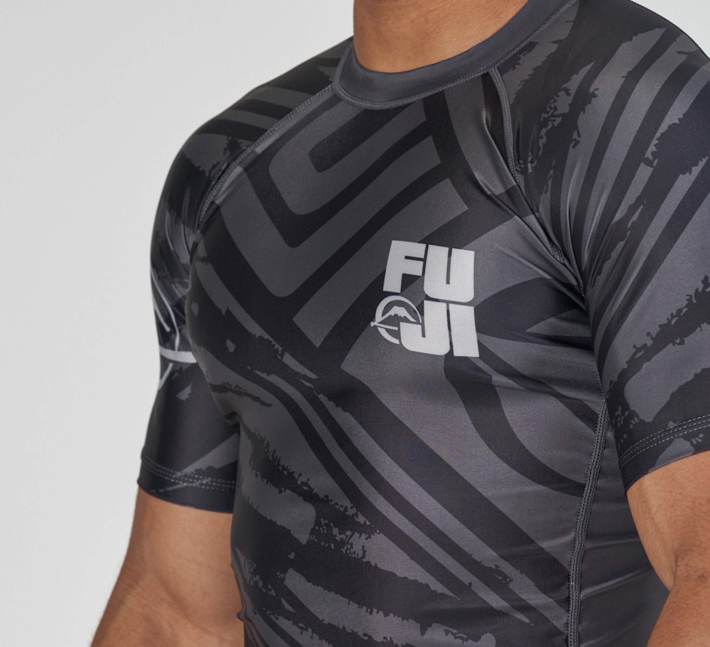 A person wearing the Battle Flex Lite Rashguard Black by FUJI, featuring abstract black and gray patterns, shown from the shoulders to upper chest and arm.