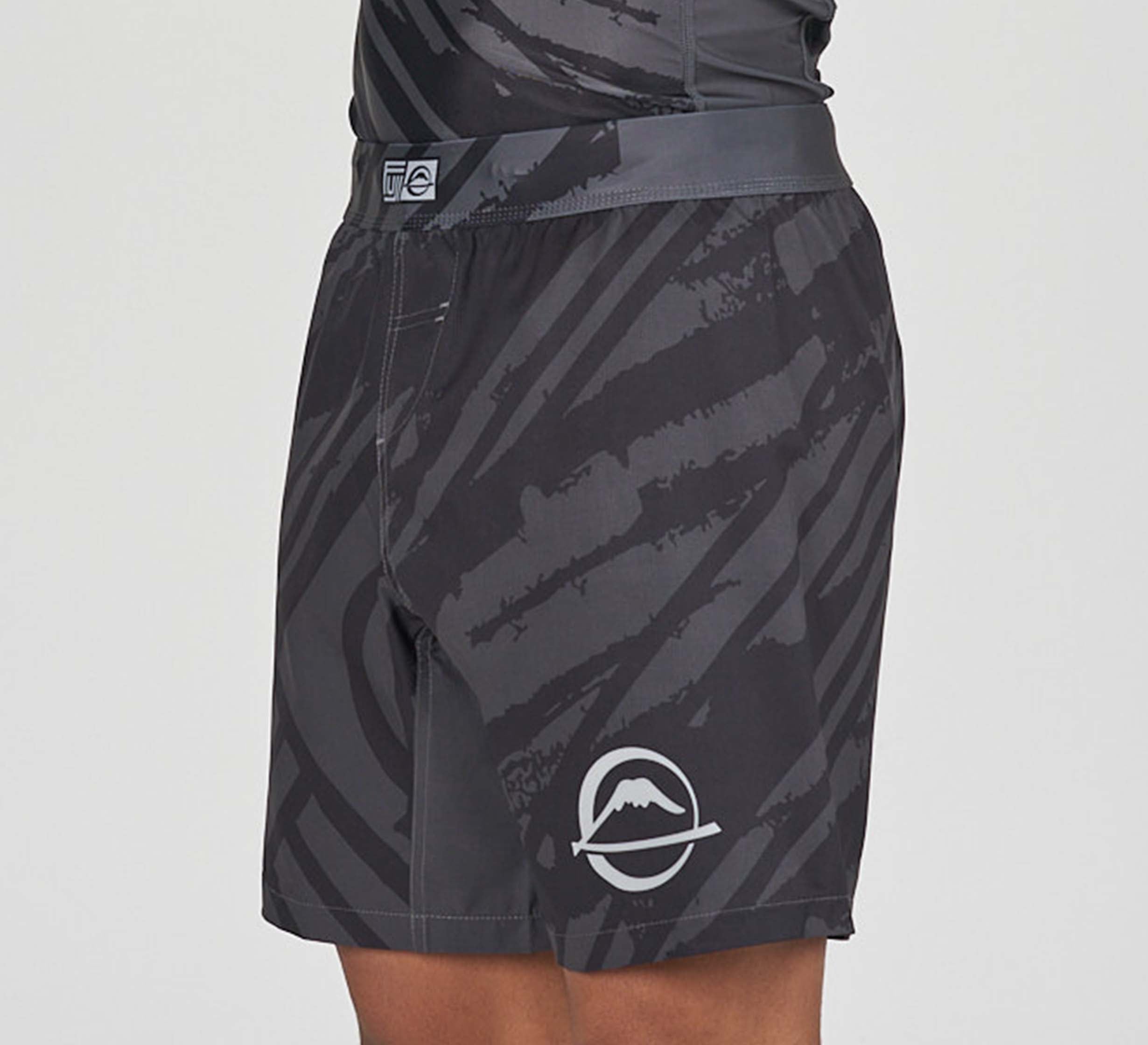 A person wearing Battle Flex Lite Shorts Black—black and gray athletic shorts with abstract patterns and a mountain-wave logo on the left leg. The upper part of a matching top is partially visible.