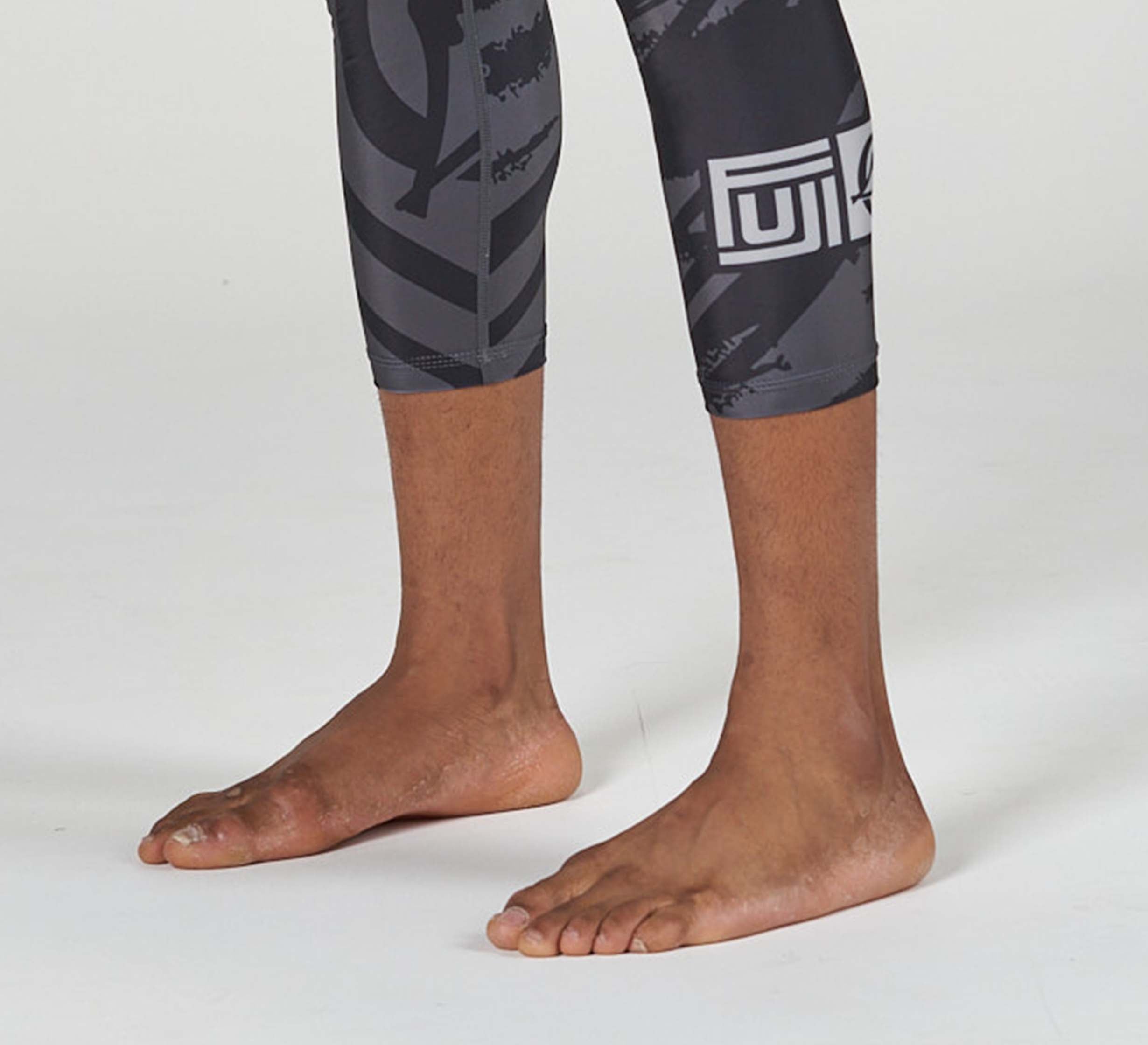 A person standing barefoot on a white surface, shown from mid-calf down, wearing Battle Flex Lite Spat Black athletic leggings with black and gray abstract patterns and logos.