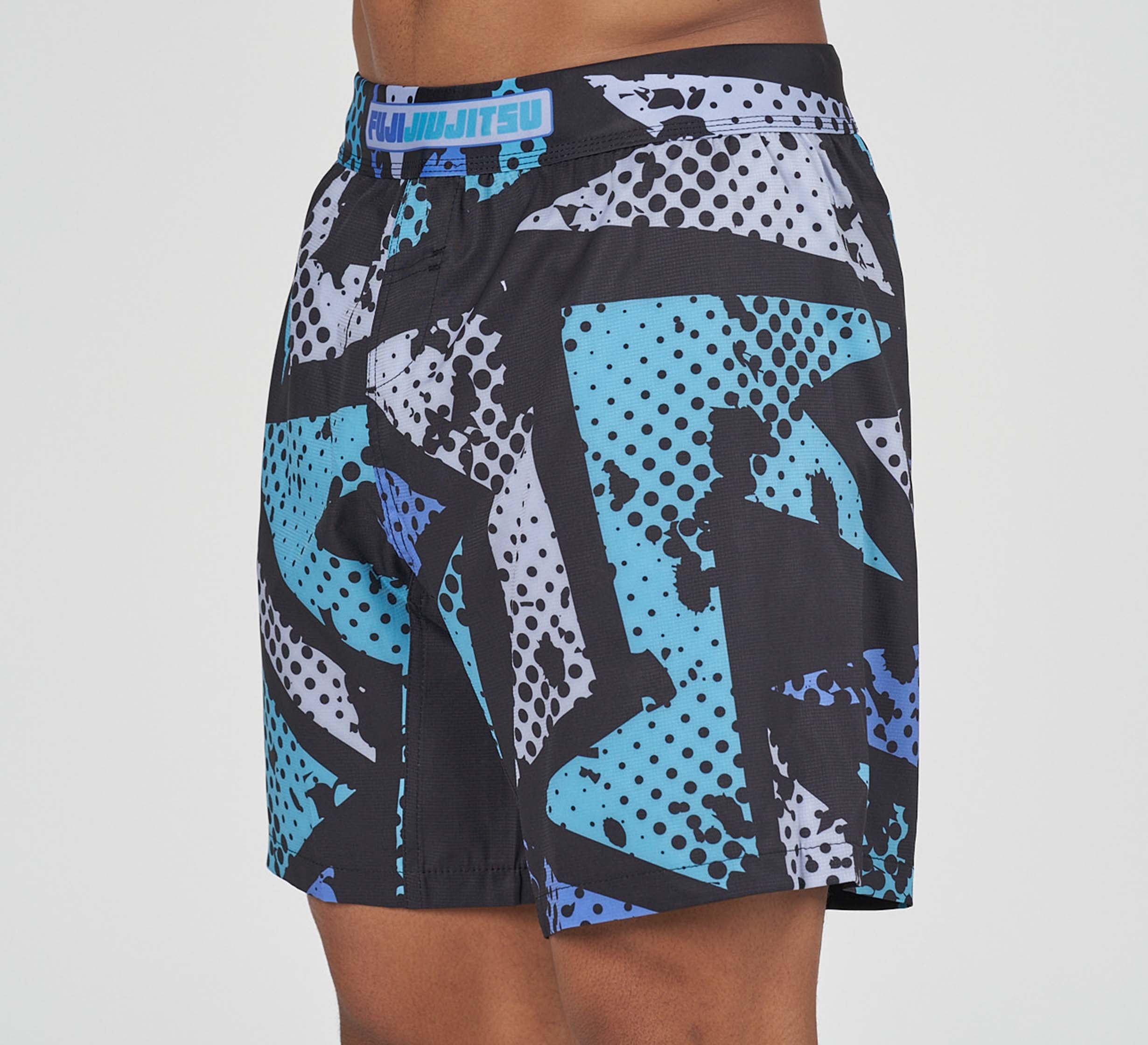 A person wears Shuriken Flex Lite Shorts Blue/Purple with a blue and white graffiti design and "Jiu Jitsu" on the waistband. The image shows only the torso and upper legs.