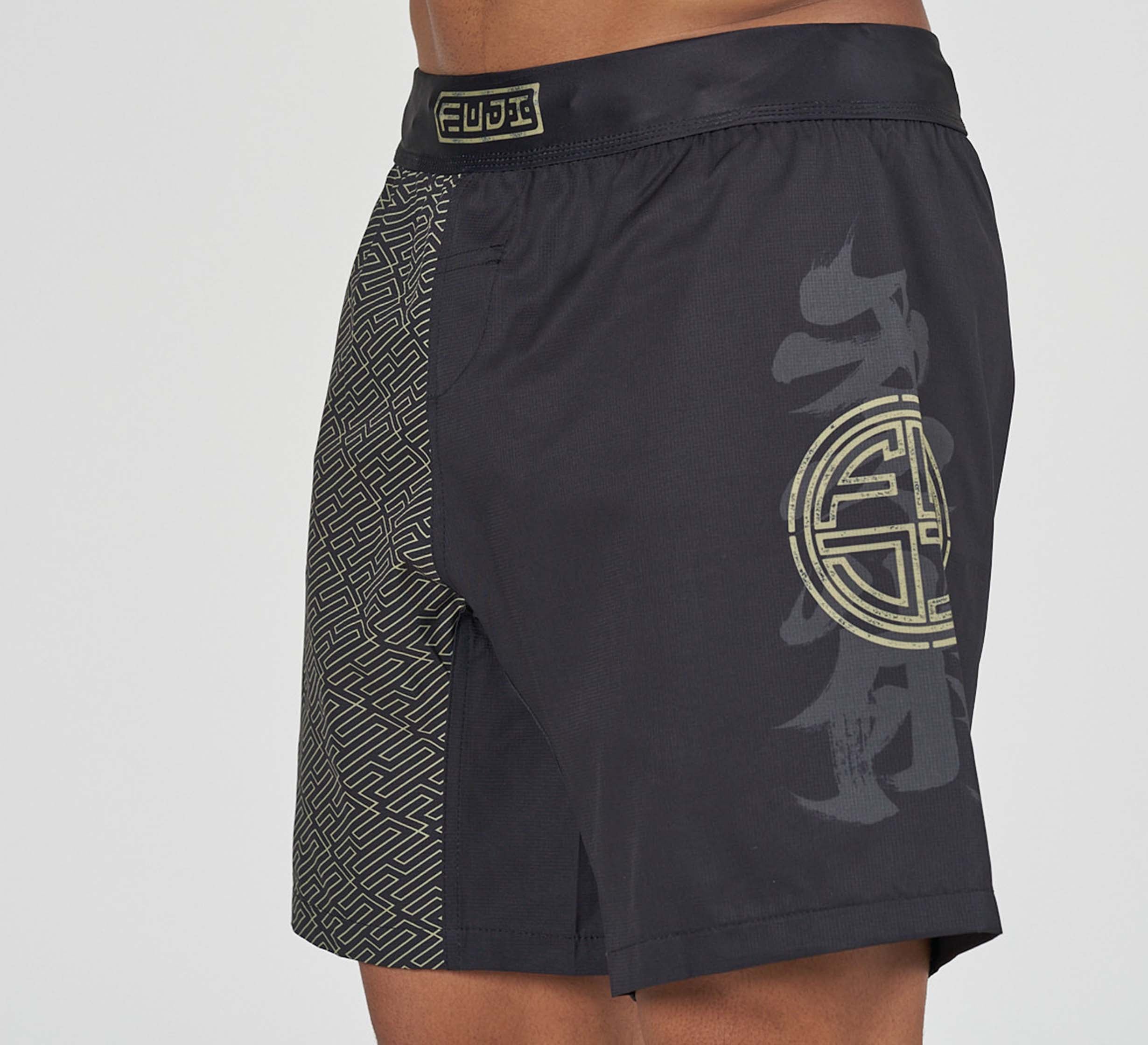 A person wearing Bushido Flex Lite Shorts, featuring black fabric, a gold geometric pattern on one side, a large gold Asian-inspired emblem on the other side, and a gold logo on the waistband.