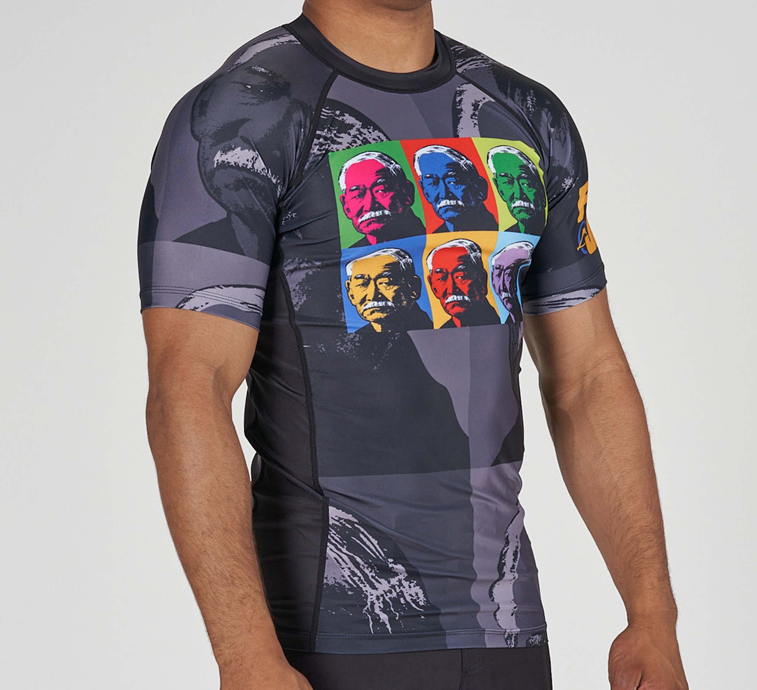 A person wears the Kano Flex Lite Rashguard, a black and gray short-sleeve athletic shirt with a vibrant pop art print of multiple faces on the front and larger, faint face images in the background.