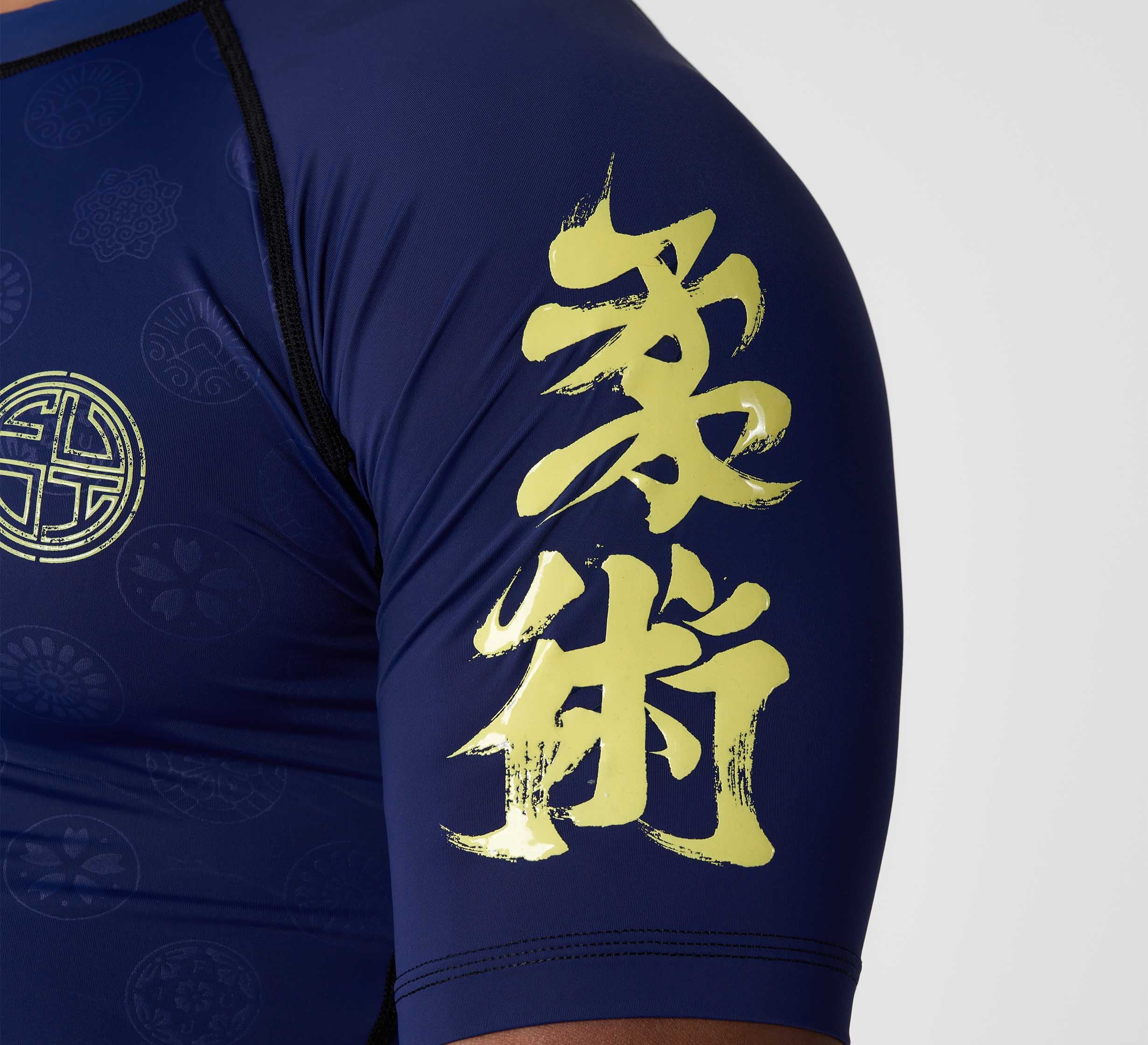 The image features a close-up of a person’s upper arm wearing the Shogun Heat Gear Rashguard Navy, which has yellow Japanese kanji characters and circular patterned designs printed on the sleeve.