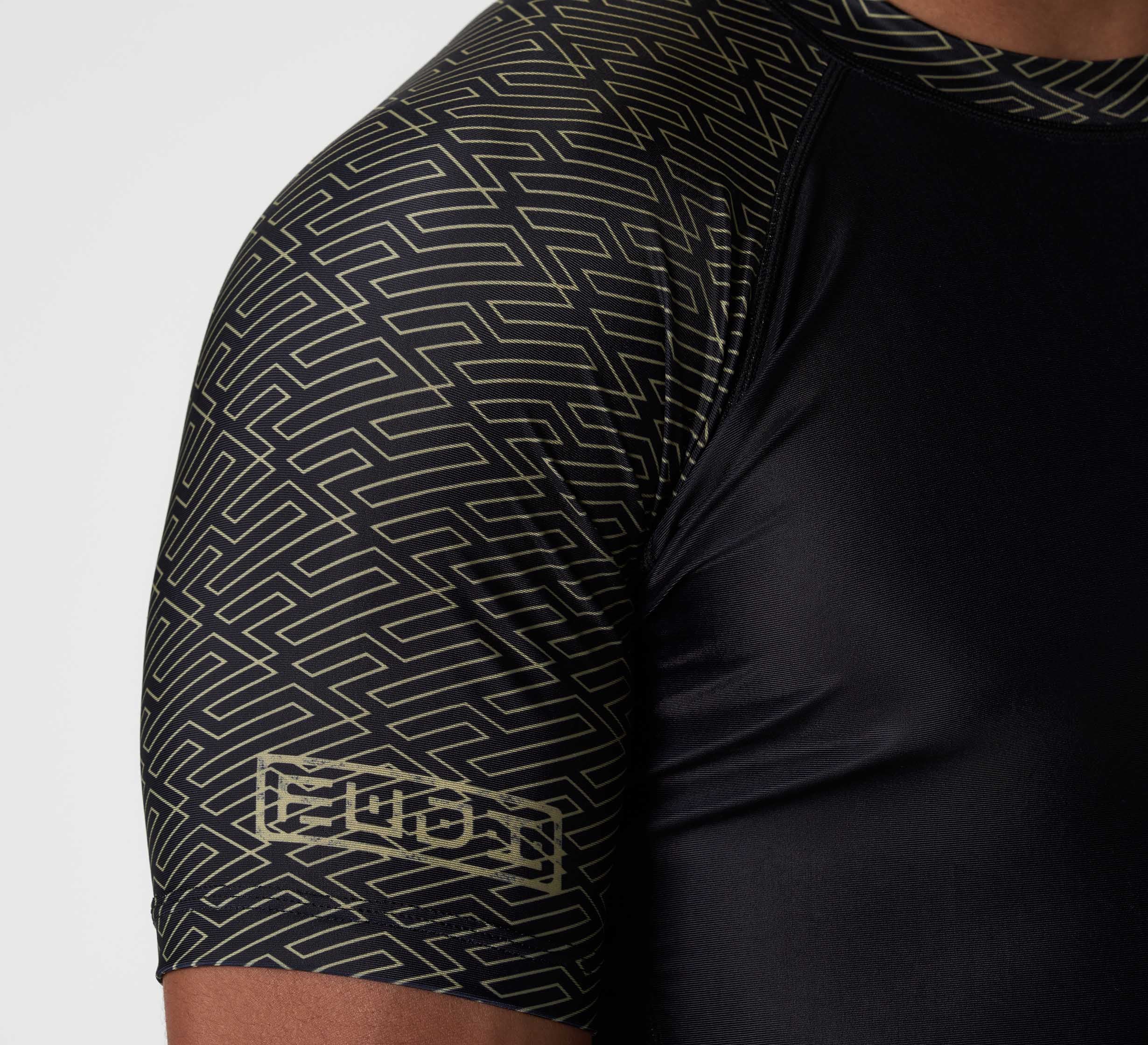 A close-up of a person wearing the Bushido Flex Lite Rashguard, a black short-sleeve athletic shirt featuring gold geometric patterns on the sleeves and shoulder, made from a smooth, stretchy material.