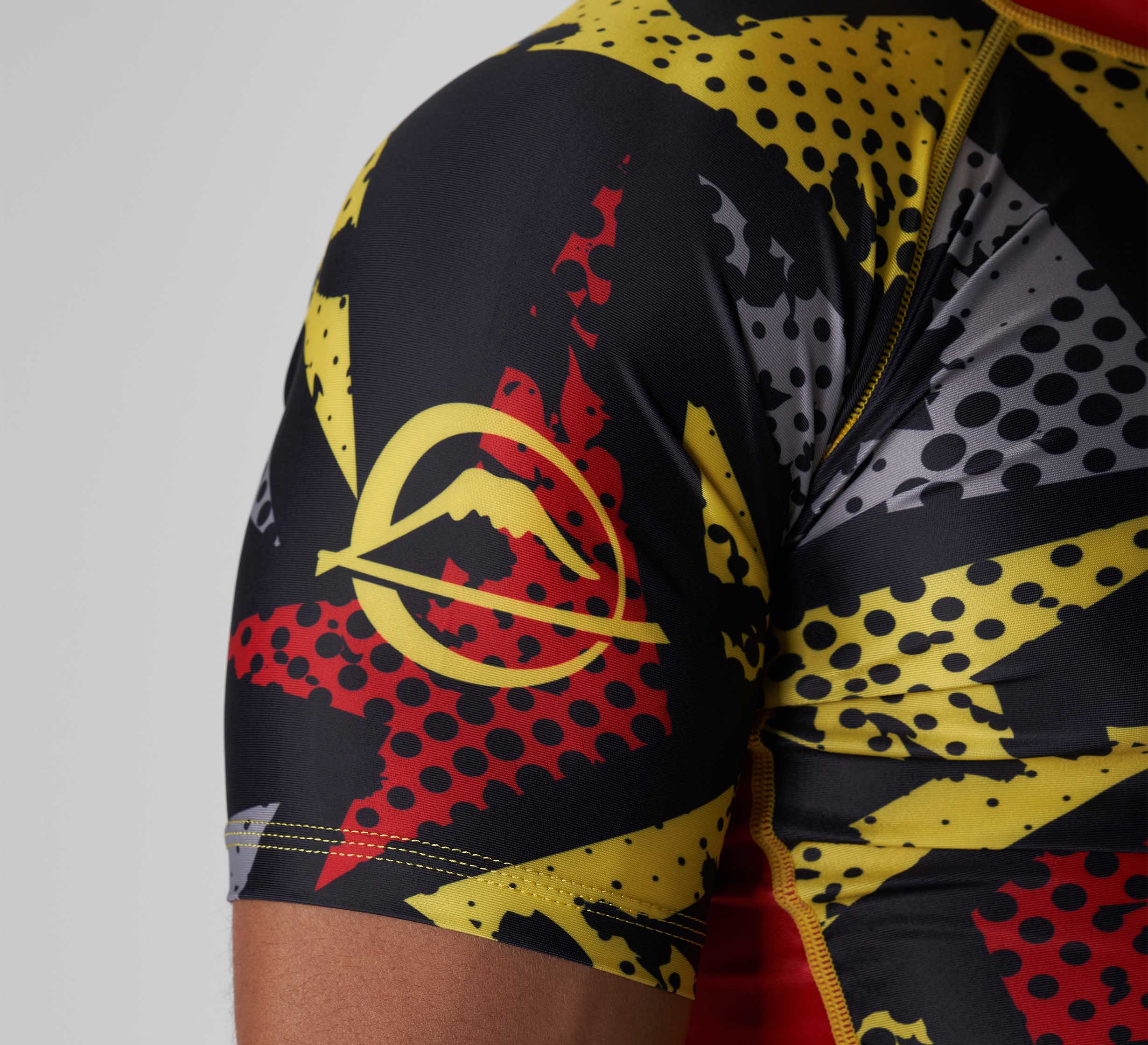 A close-up of a person's upper arm wearing the Shuriken Flex Lite Rashguard Black/Yellow, featuring a bold red, yellow, and black comic-style pattern and a yellow circular logo with a mountain silhouette.
