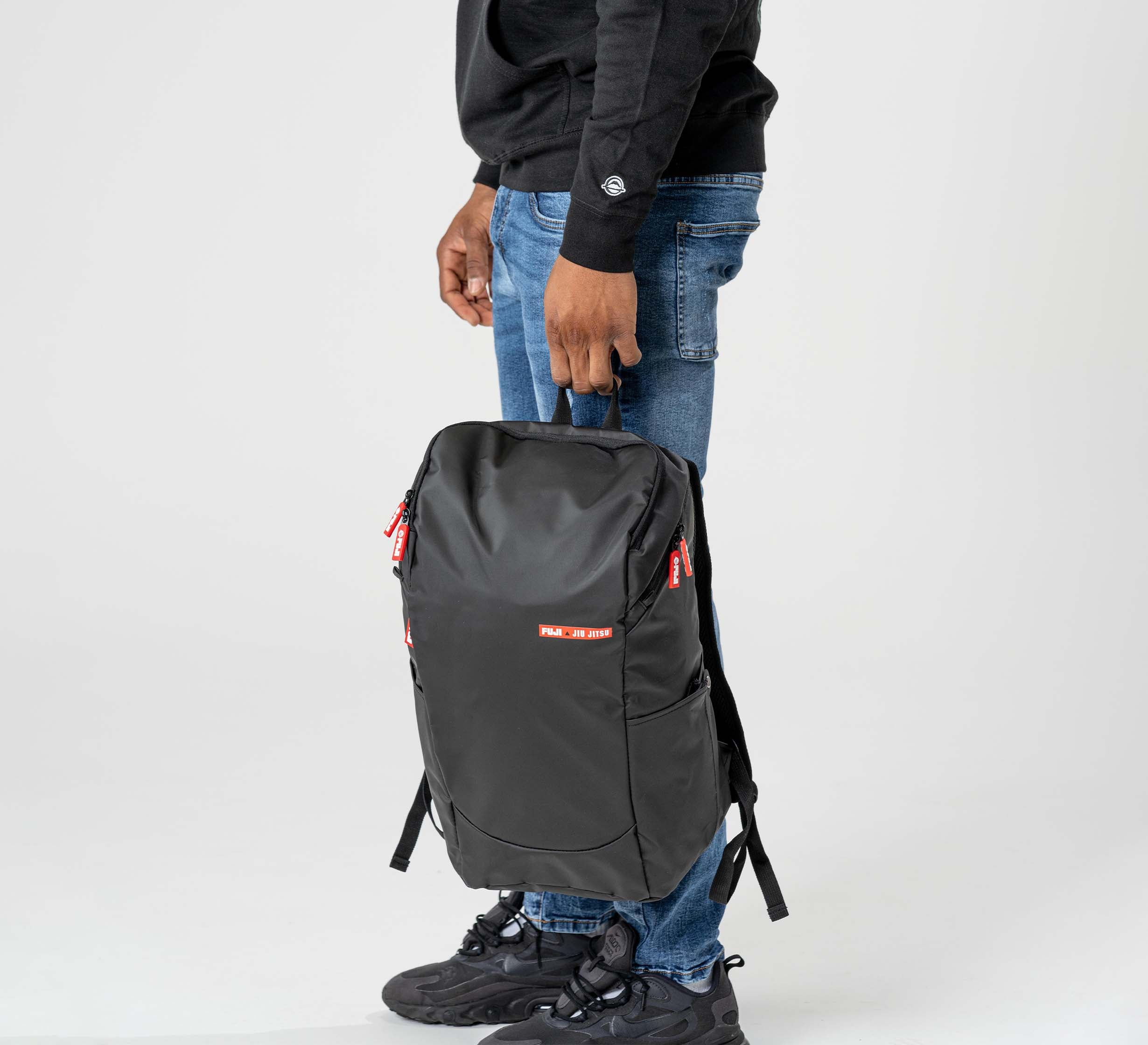 Someone in a black sweatshirt, blue jeans, and black sneakers stands holding the Urban Day Backpack Black by its top handle against a plain light background. The backpack has red zipper accents and a small rectangular label.