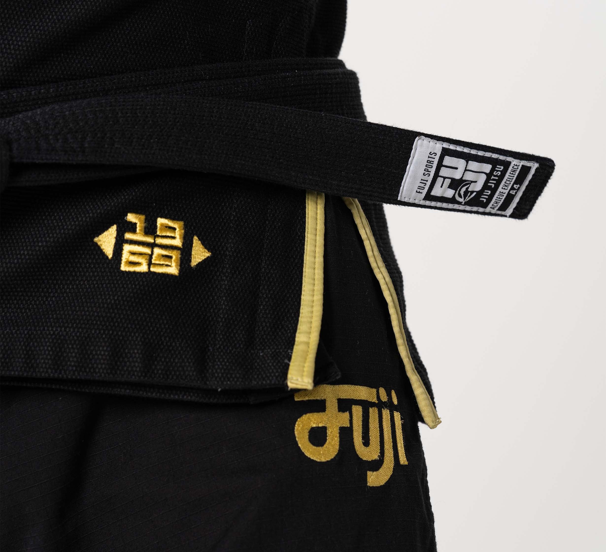 Close-up of a person wearing the Flow-Tech BJJ Gi Black/Gold, featuring gold embroidery, a tied black belt, and the FUJI Sports brand label.