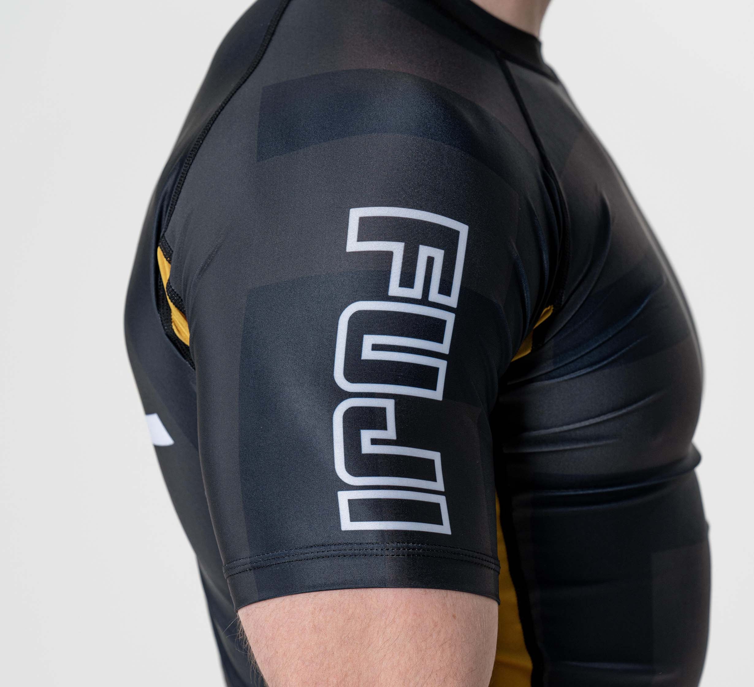A person wearing the Rebel Flex Lite Rashguard Black, a tight athletic shirt with "FUJI" in large white letters on the sleeve, featuring yellow and gray accents.