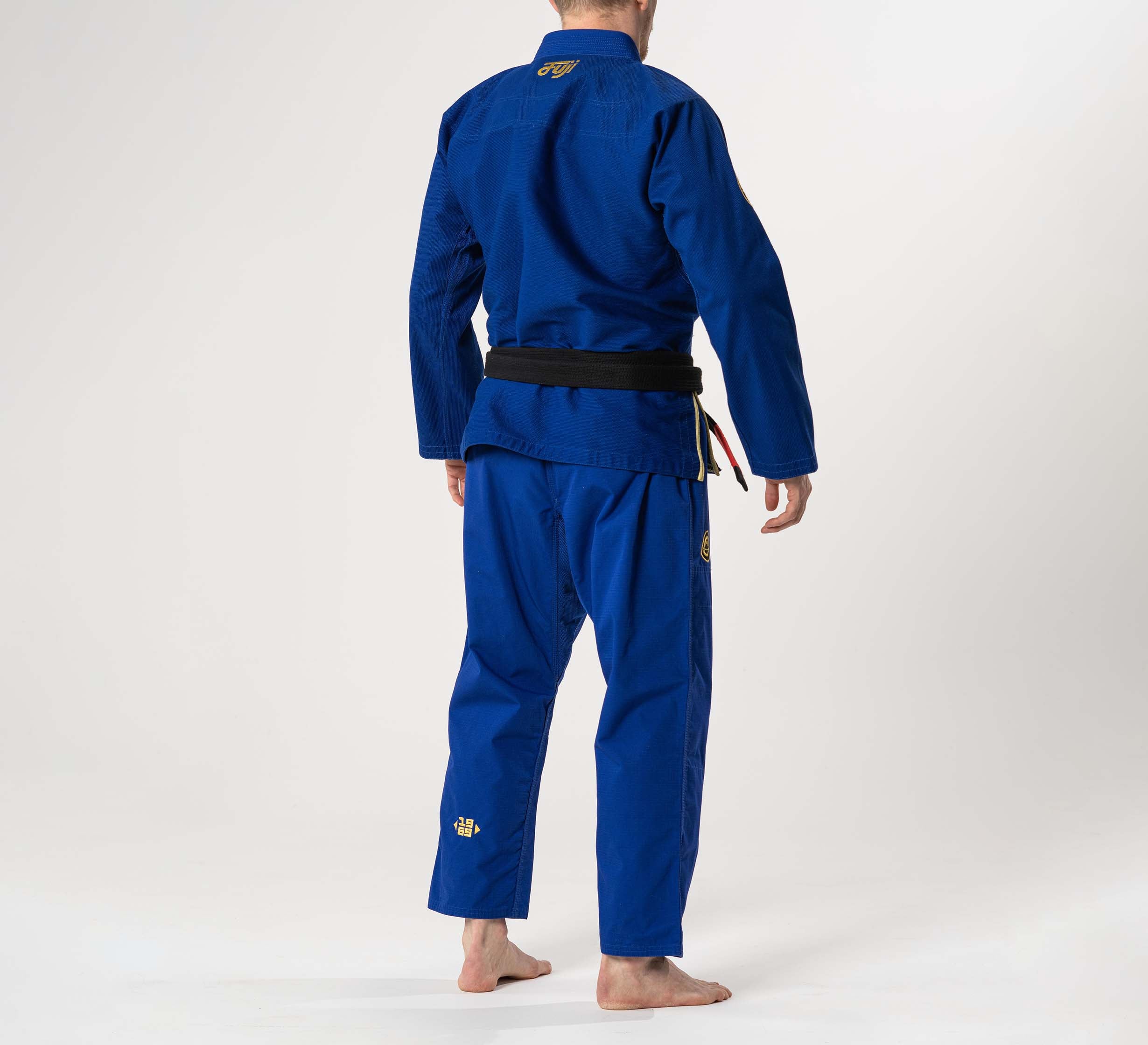 A barefoot person wearing the Flow-Tech BJJ Gi Blue/Gold with a black belt stands facing away from the camera against a plain, light background.