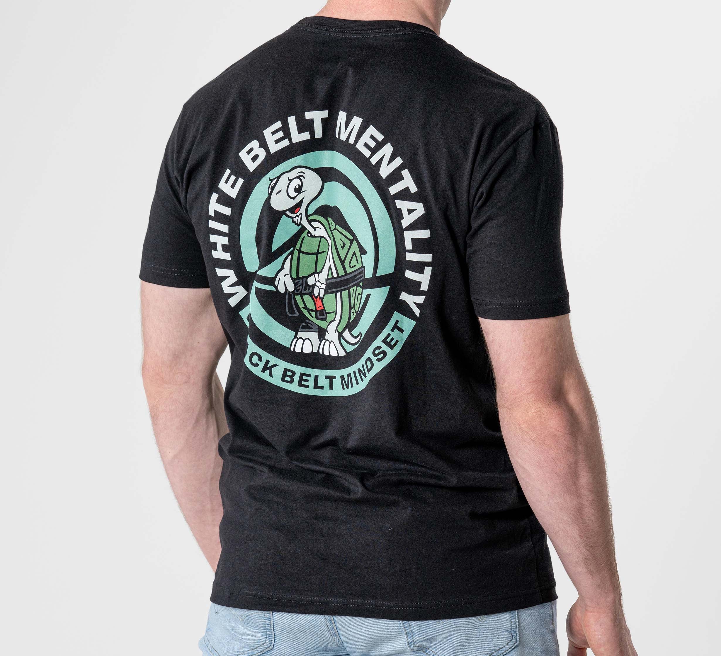 A man wearing the Black Belt Mentality T-Shirt Black, featuring a cartoon turtle holding a black belt on the back. The shirt says “WHITE BELT MENTALITY” above and “BLACK BELT MINDSET” below the illustration.