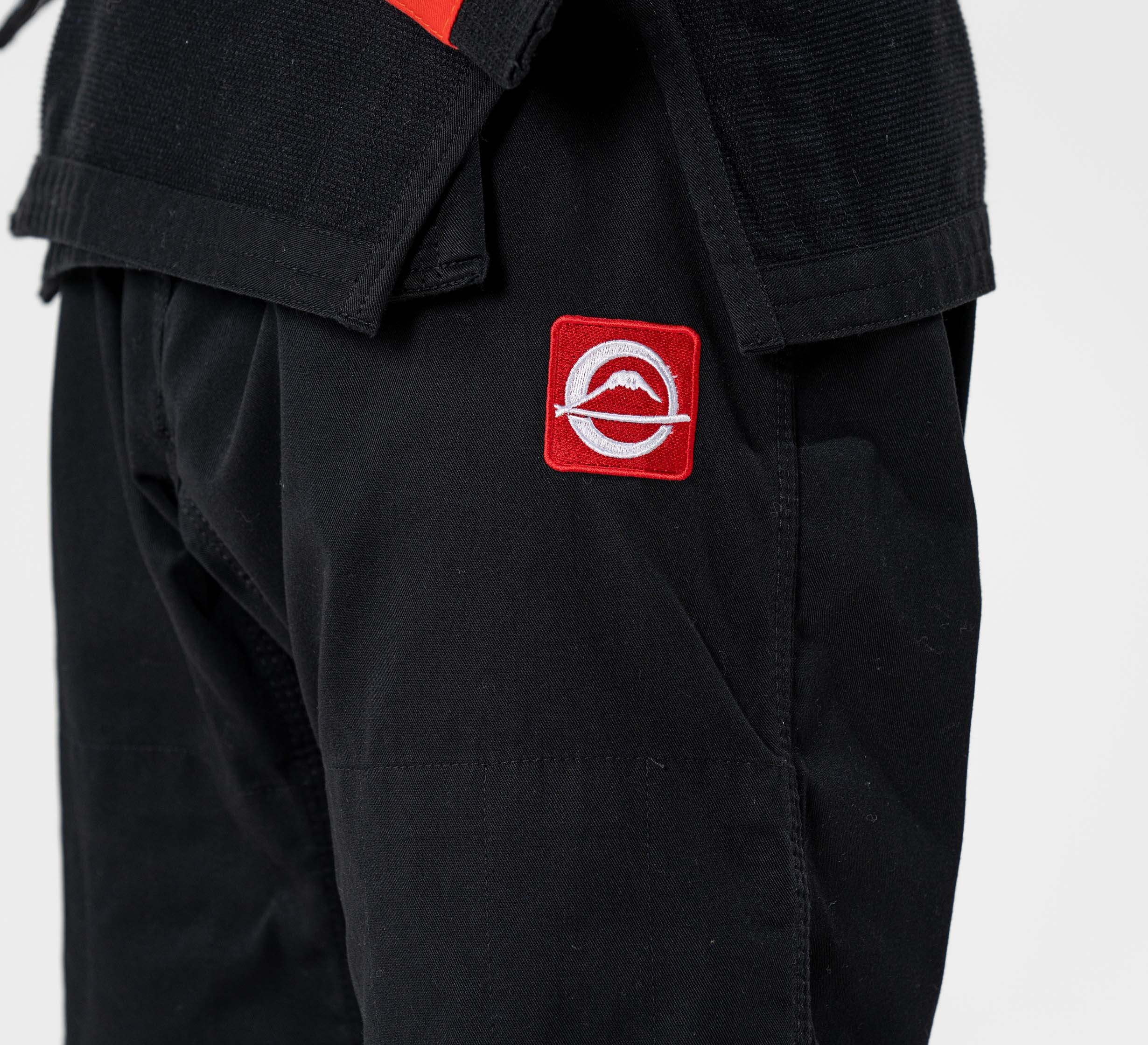 Close-up of a person wearing the Kids Ultra Lite 300 BJJ Gi Black, featuring black martial arts pants with a red and white circular patch on the thigh showing a stylized shark fin logo crossed by a diagonal line.