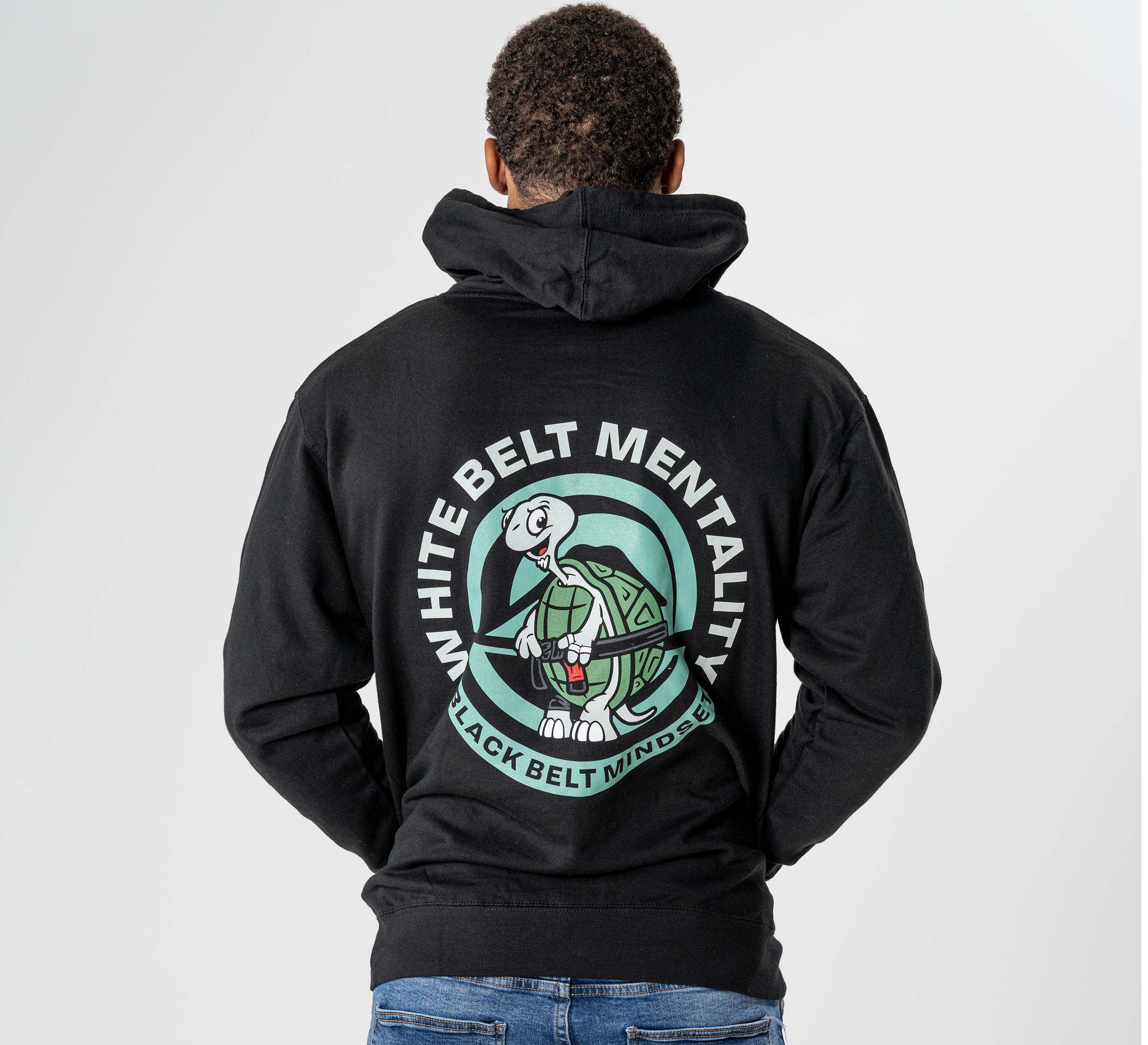 A person wearing the Black Belt Mentality Hoodie Black, featuring a cartoon turtle logo and the text "WHITE BELT MENTALITY BLACK BELT MINDSET" on the back, stands against a plain light background.