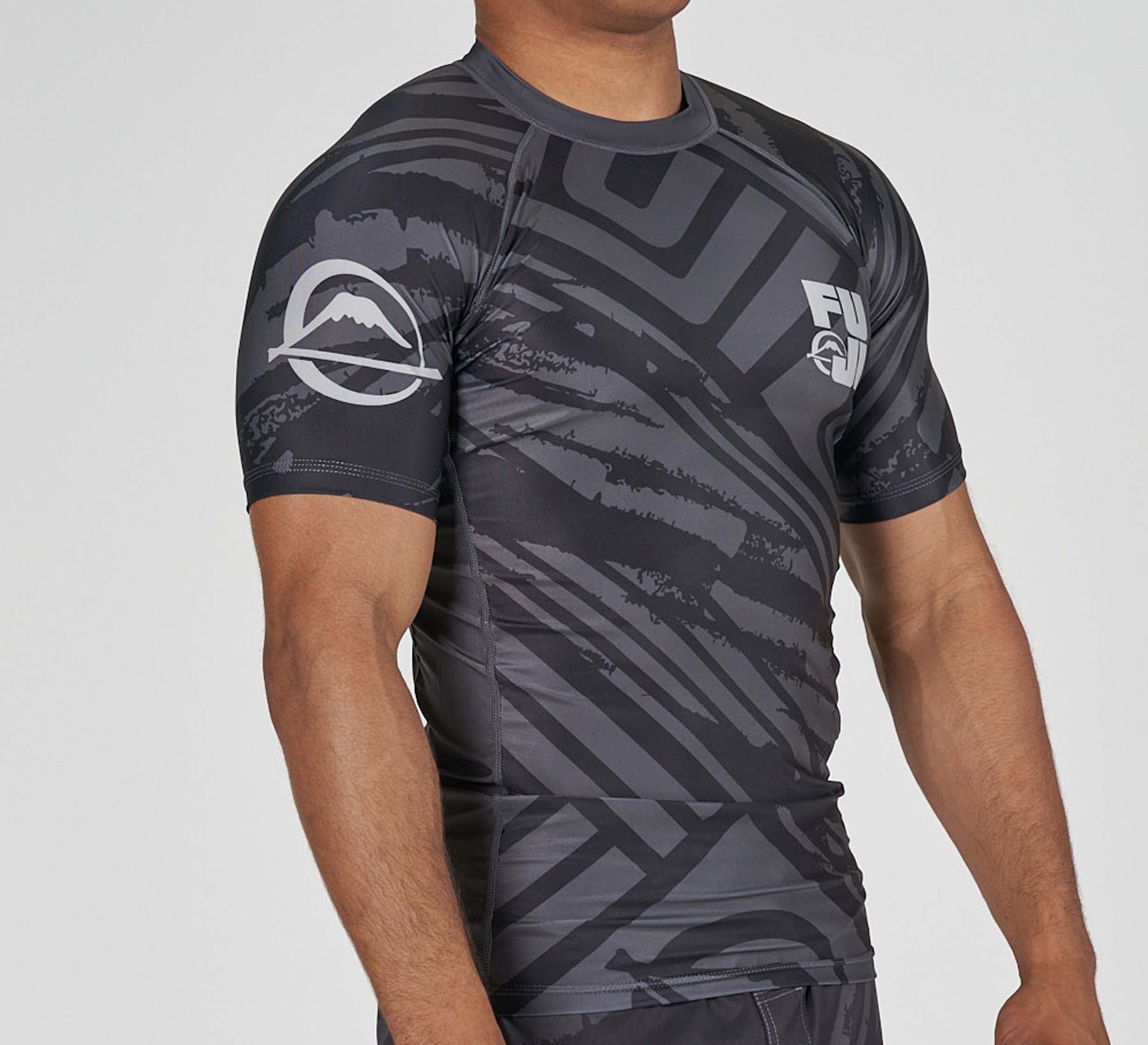 A person wears the Battle Flex Lite Rashguard Black, a fitted short-sleeve top featuring abstract black and gray patterns with white logos on the chest and sleeve, standing against a plain background.