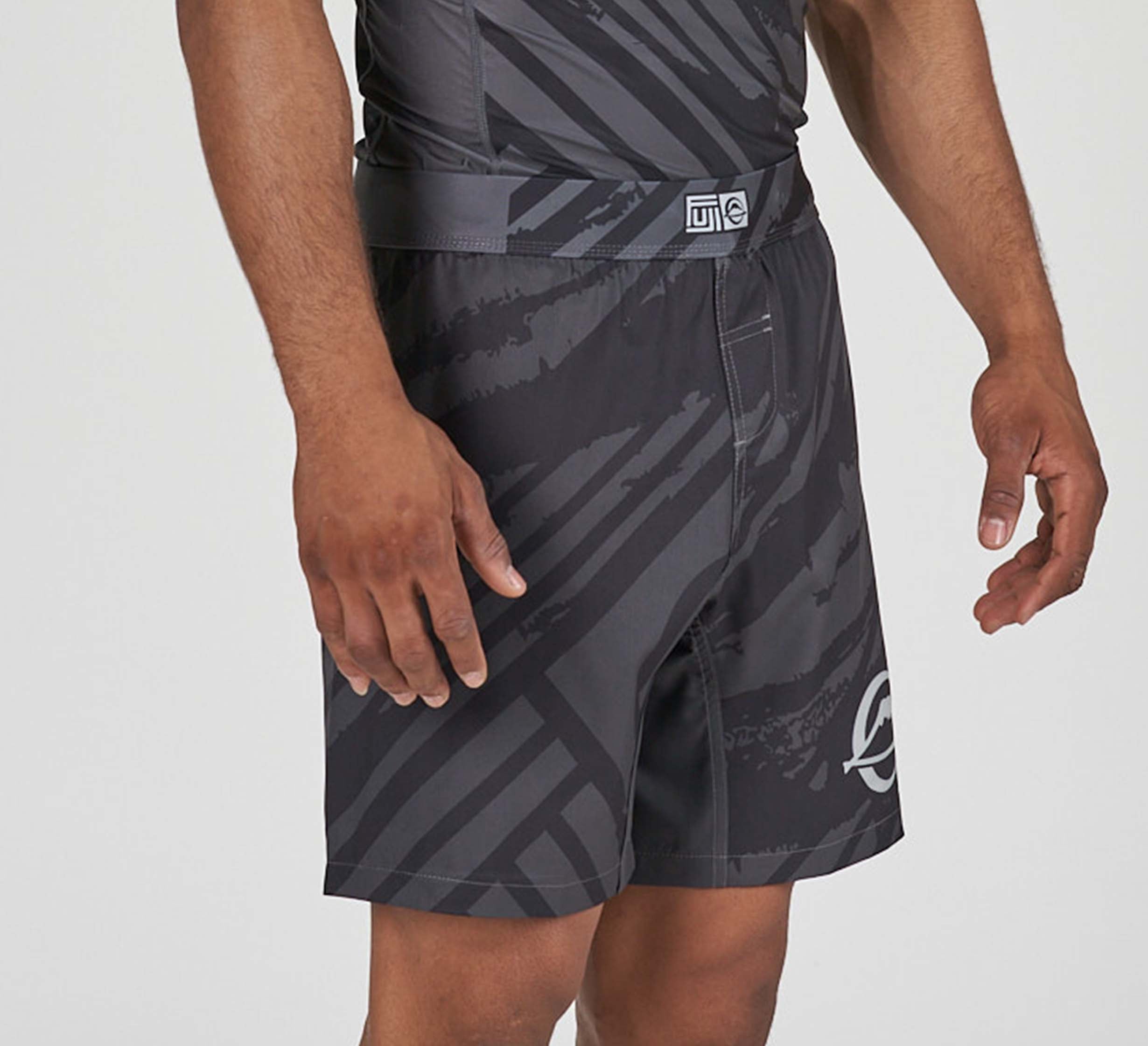 A person wears Battle Flex Lite Shorts Black featuring a black and gray pattern, a belt, and a white circular logo on the left leg. One arm is relaxed at their side while the other is slightly bent.