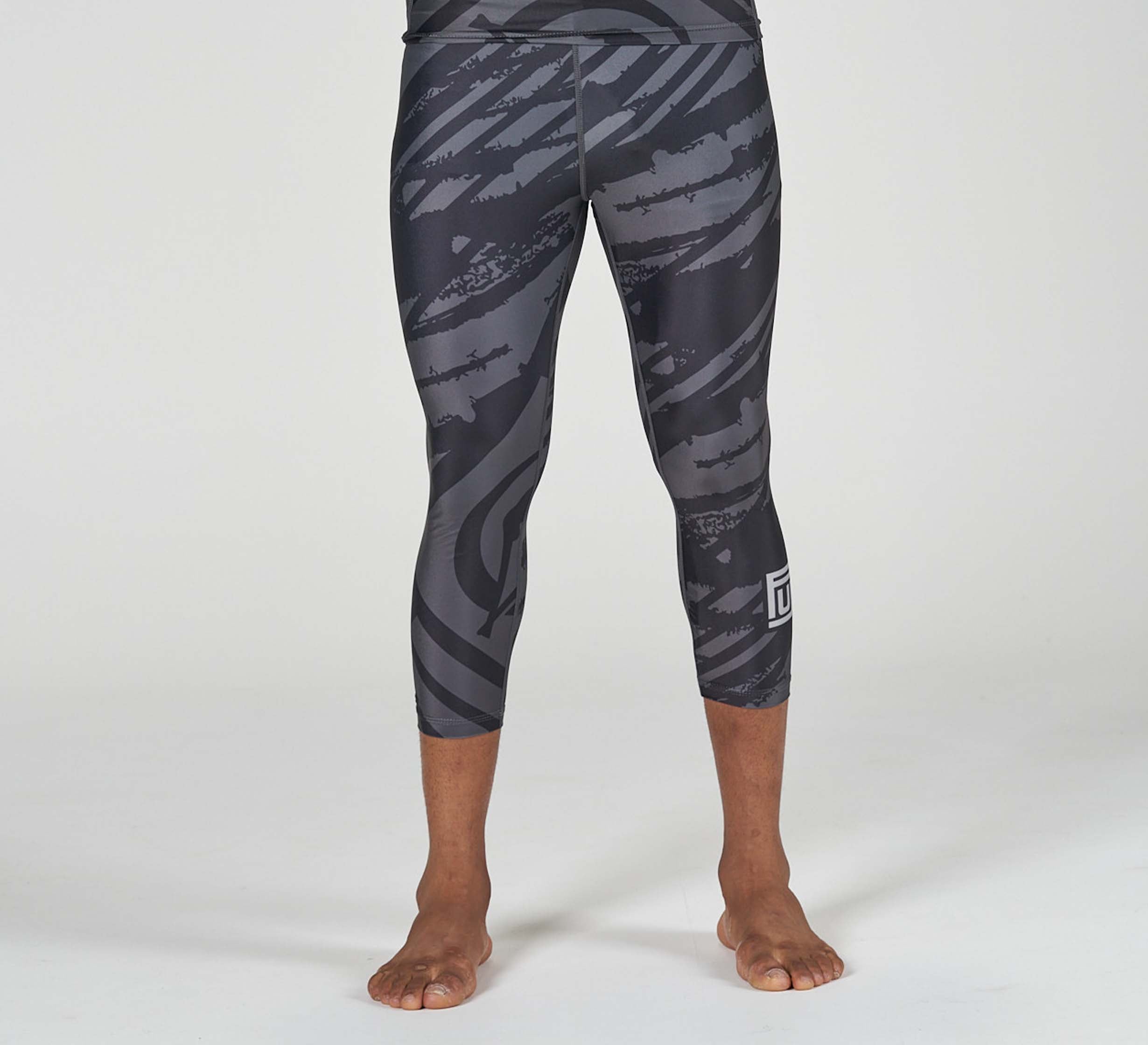 A person stands barefoot against a plain light background, wearing Battle Flex Lite Spat Black compression leggings featuring a black and gray pattern with a small white logo on the left calf.