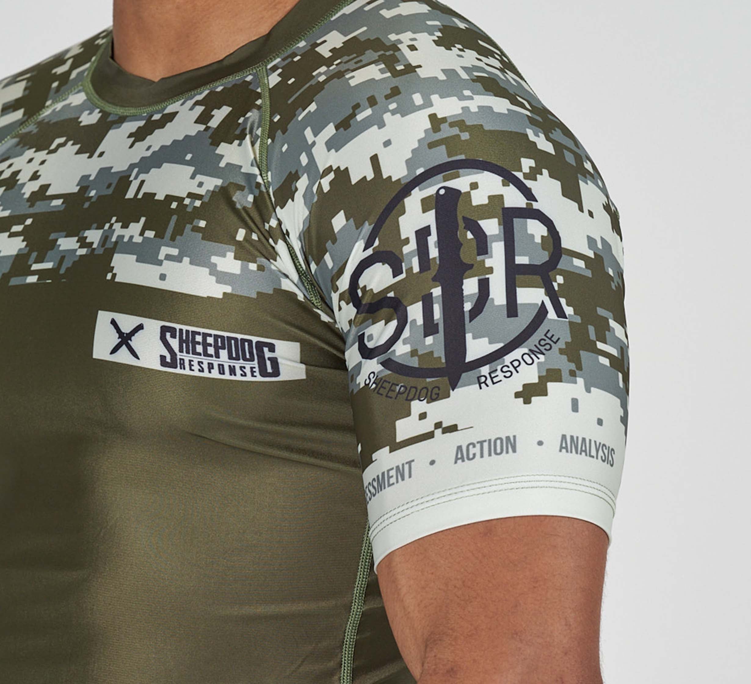 A person wearing the Sheepdog Response Flex Lite Rashguard Military Green, featuring Sheepdog Response patches on the chest and sleeve, and “ASSESSMENT - ACTION - ANALYSIS” printed on the armband.