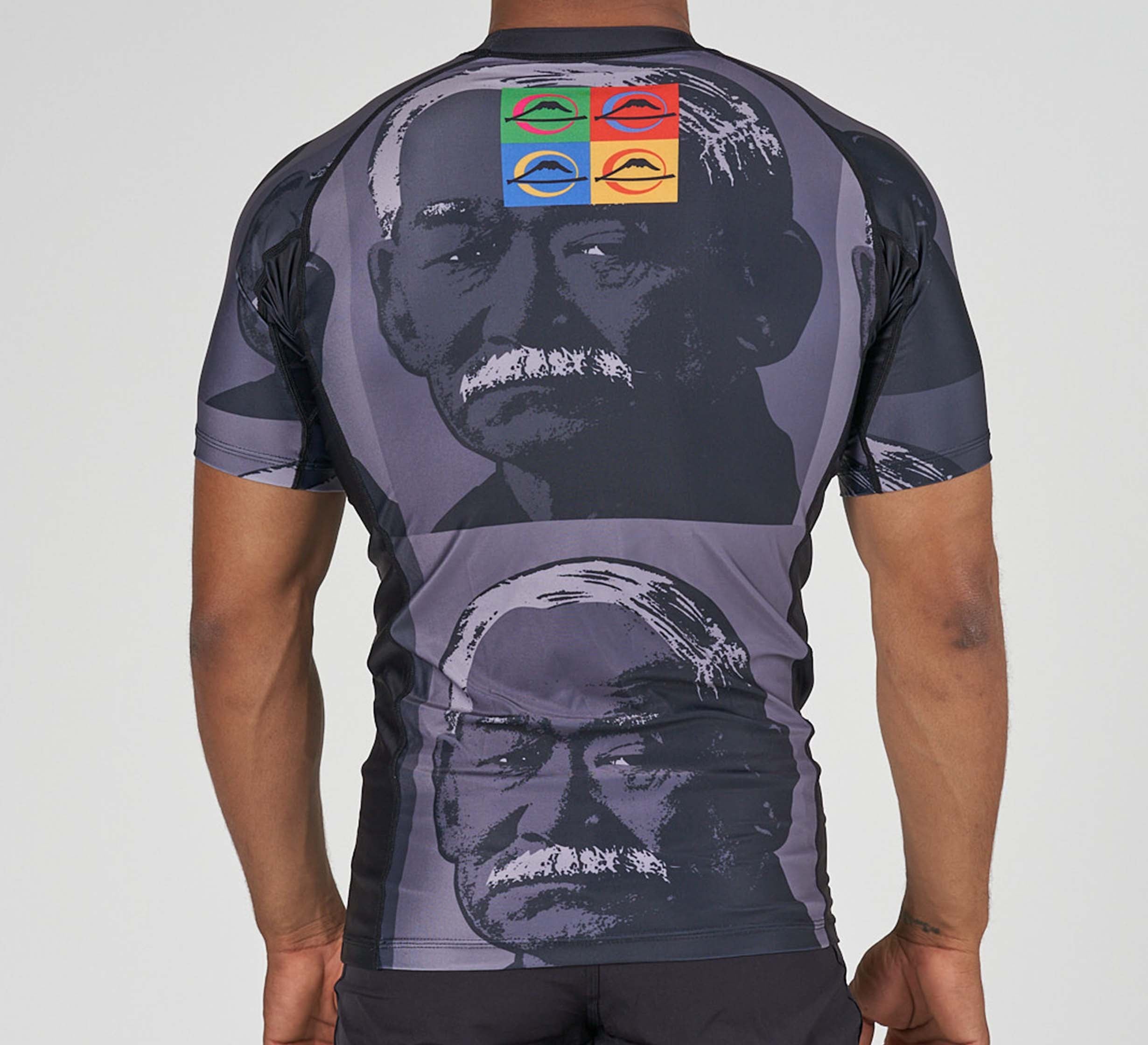 Someone wearing the Kano Flex Lite Rashguard, a short-sleeved shirt with grayscale portraits of an older man with a mustache and four colorful eye symbols—red, orange, green, and blue—on the upper back.