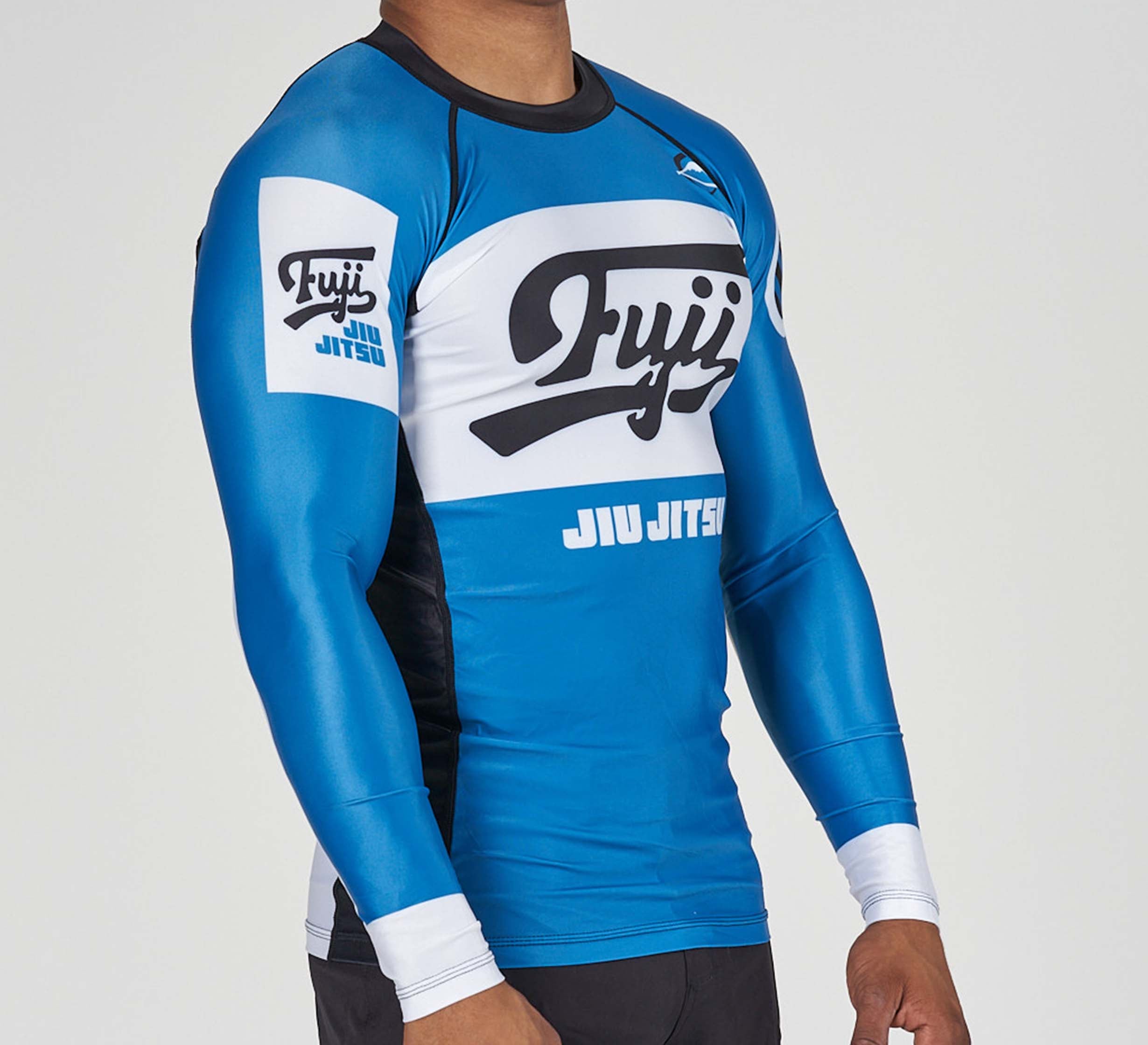 Someone wearing the Jiu Jitsu Script Flex Lite Long Sleeve Rashguard in blue, featuring Fuji and JIU JITSU logos on the chest and sleeve, stands against a plain background.