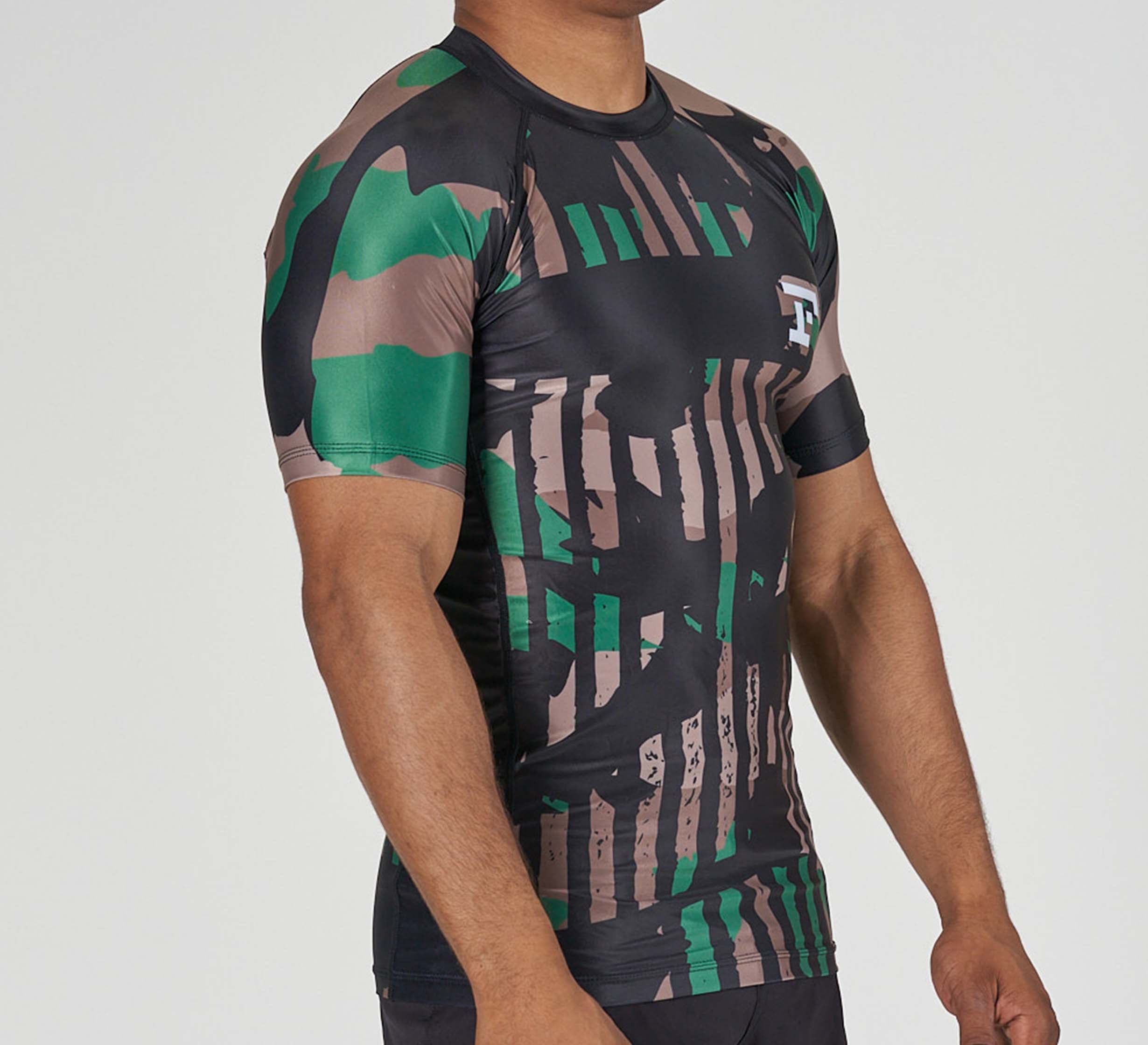 Someone stands against a plain background wearing the Camo Flex Lite Rashguard, a fitted short-sleeve athletic shirt featuring a green, brown, and black camouflage pattern.