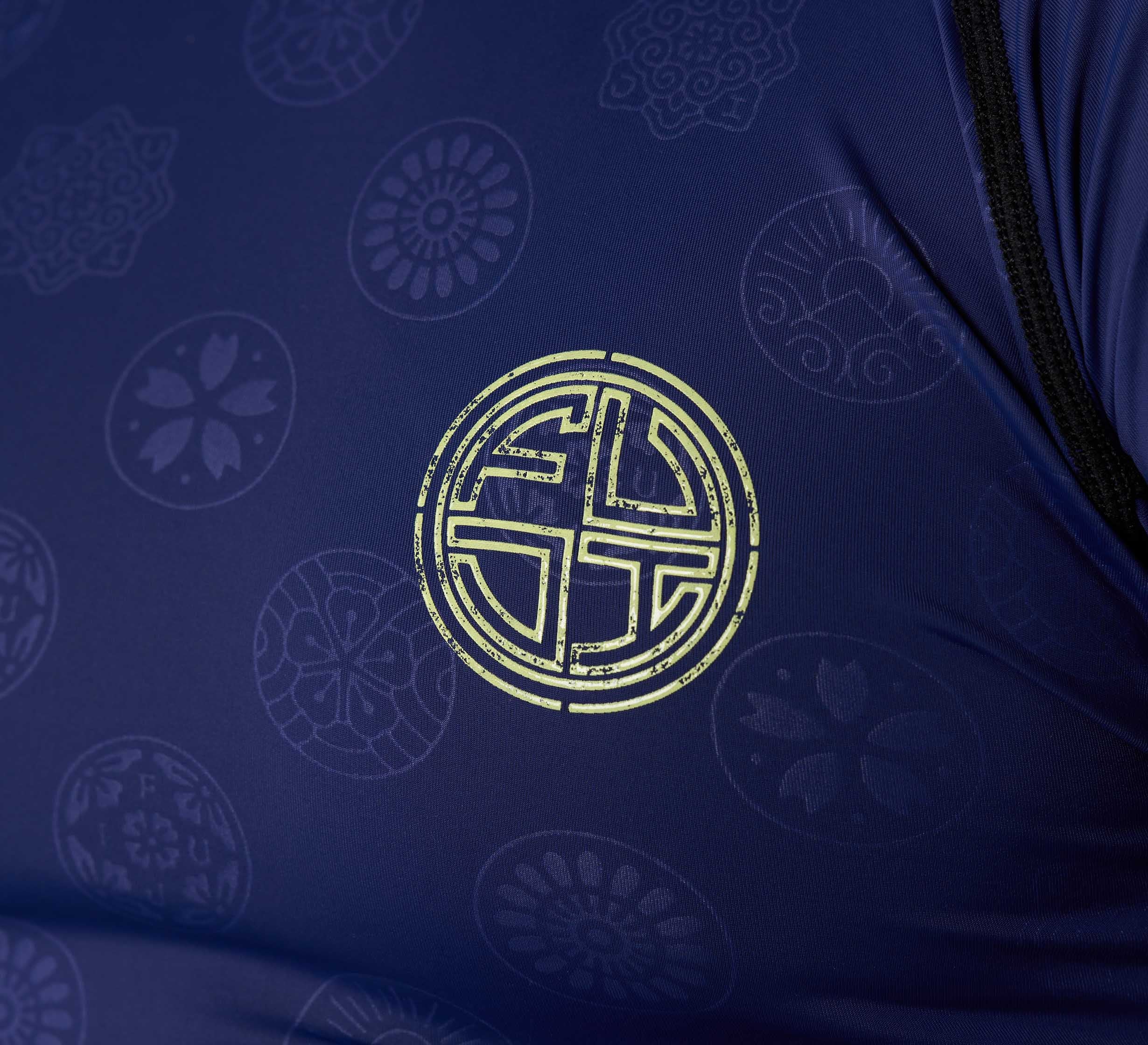 Close-up of the Shogun Heat Gear Rashguard Navy, featuring navy blue fabric with subtle floral patterns and a bold yellow circular emblem at the center. Visible black seams run along the right edge.