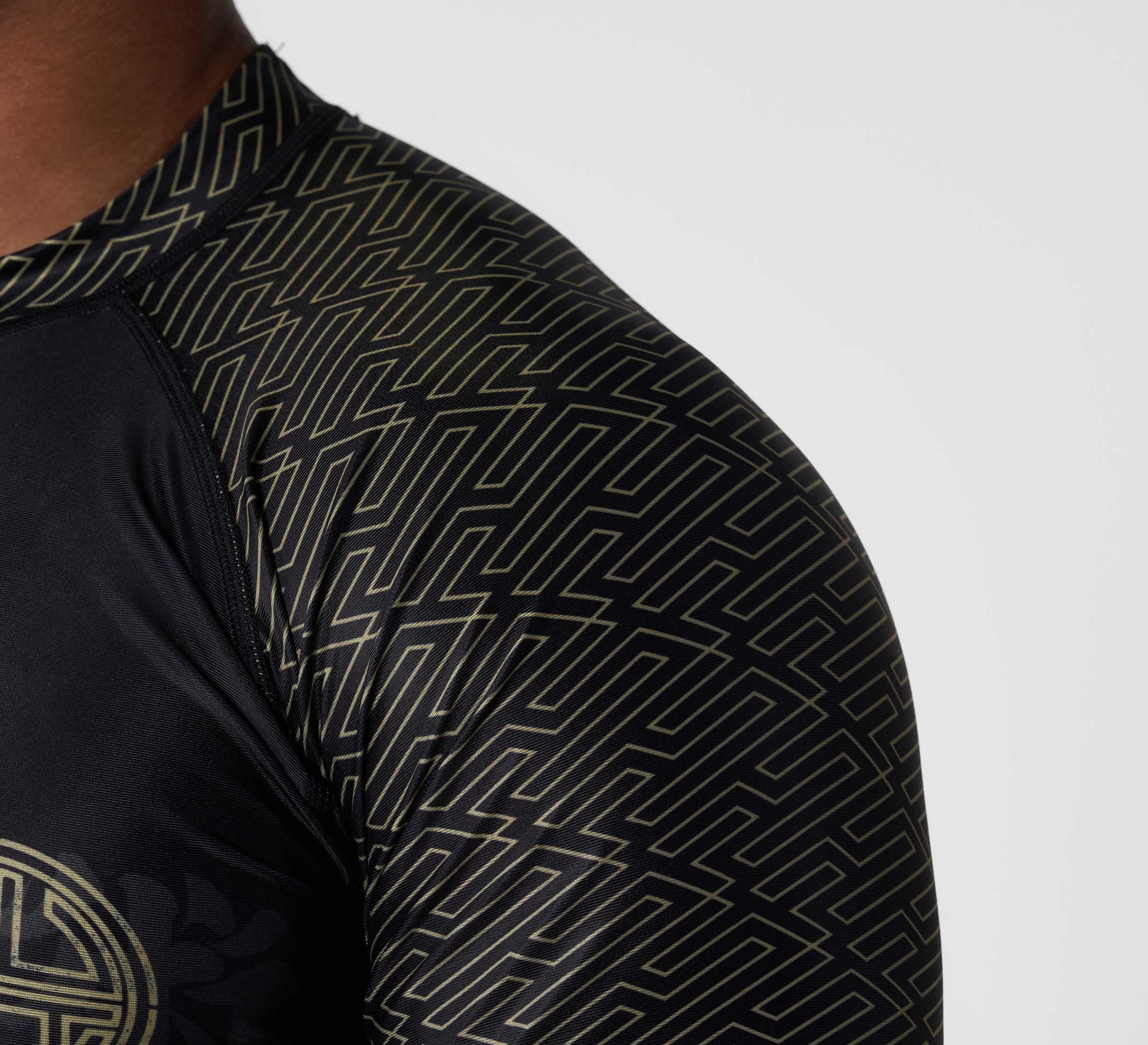 Close-up of a person wearing the Bushido Flex Lite Rashguard, a black athletic shirt featuring gold geometric maze-like patterns on the shoulder and sleeve. The background is plain and light-colored.
