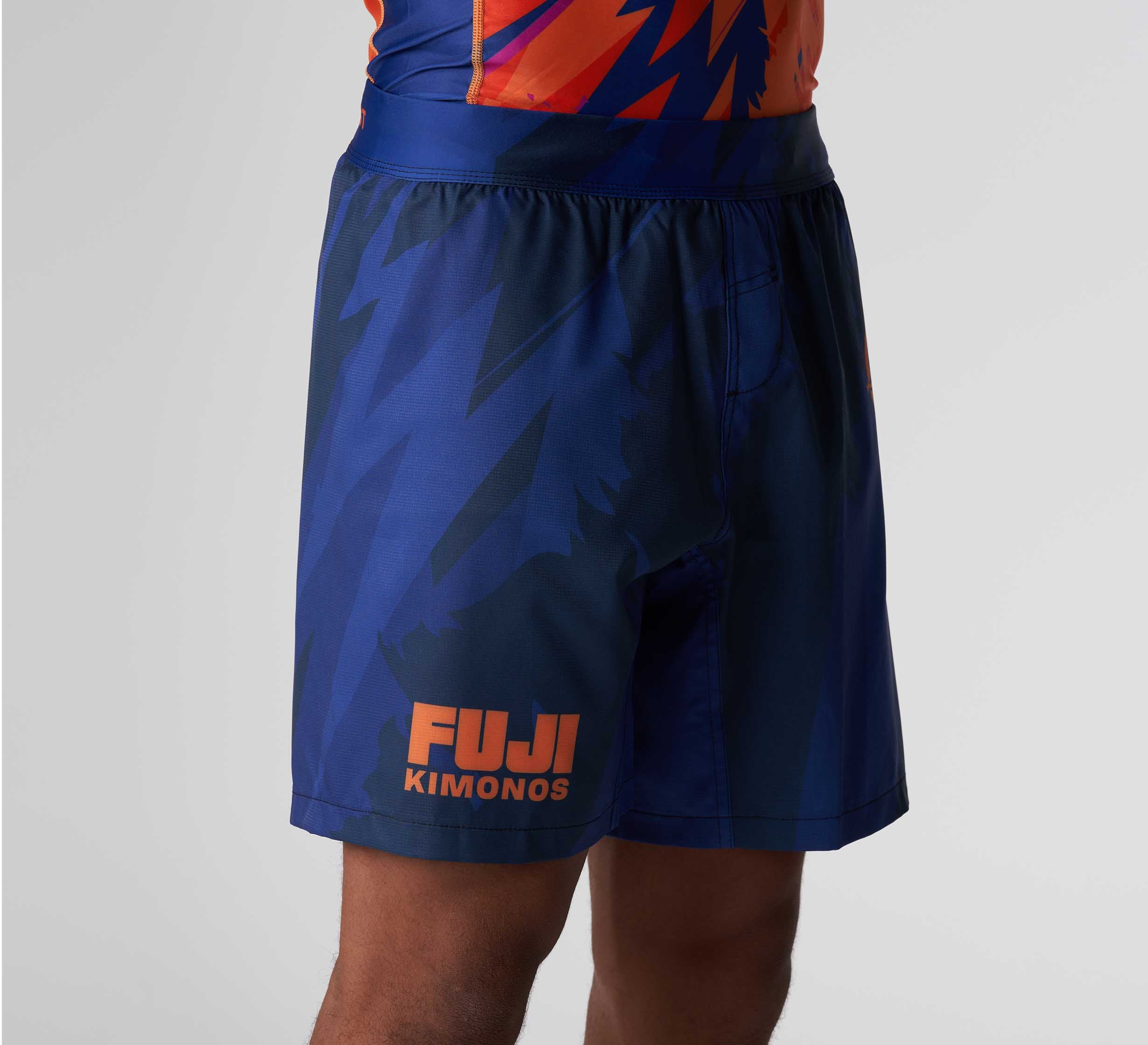 A person wearing Hanzo Flex Lite Shorts Blue featuring an abstract pattern and "FUJI Kimonos" in orange on the left leg. Only their torso and upper legs are shown against a plain background.