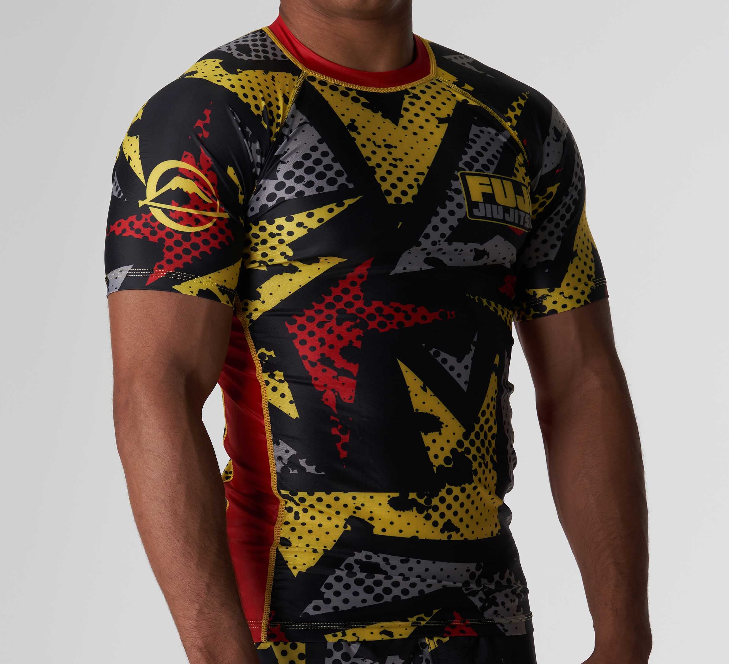 A person wearing the Shuriken Flex Lite Rashguard Black/Yellow—a short-sleeved, form-fitting rash guard featuring a bold black, yellow, gray, and red abstract design with a FUJI Jiu Jitsu logo on the chest.