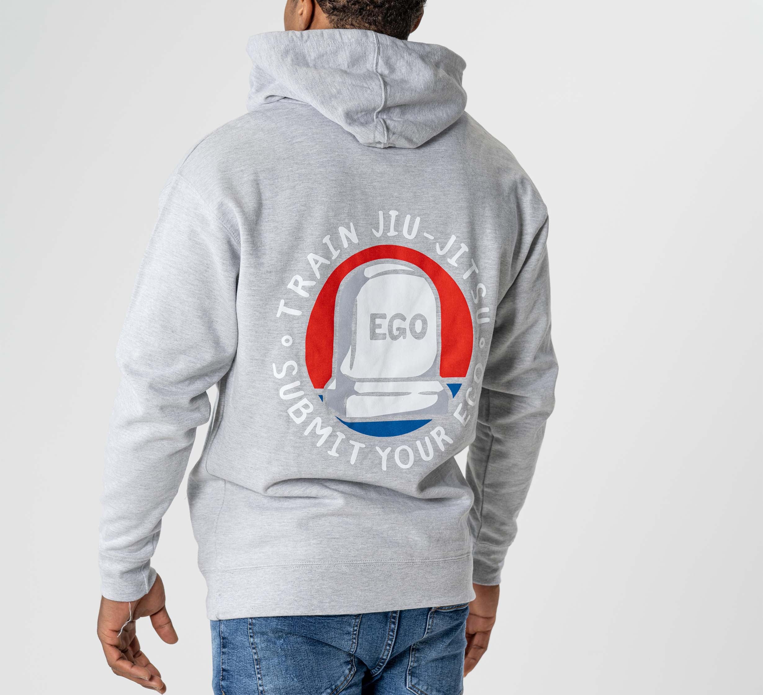 A person is seen from behind wearing the Submit Your Ego Hoodie Heather Grey, featuring a white jiu-jitsu belt graphic and "EGO" on the back. Surrounding text reads: "Train Jiu-Jitsu. Submit Your Ego.