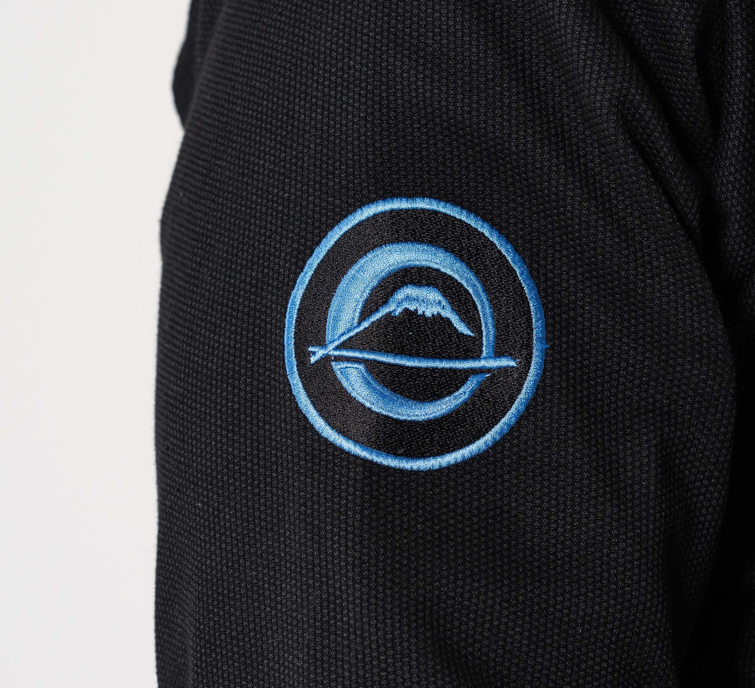 A close-up of the Flow-Tech BJJ Gi in black fabric, featuring an embroidered blue and grey circular logo with a mountain and a line passing through it.