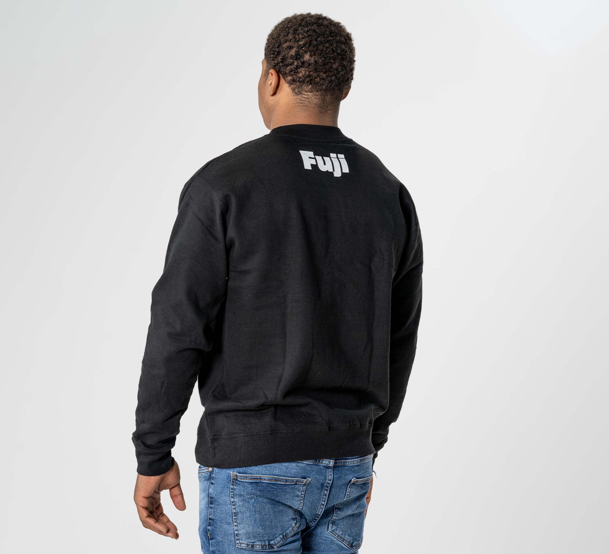 A person stands with their back to the camera, wearing the Jiu Jitsu Player Crewneck Black featuring "Fuji" in white on the upper back, paired with blue jeans against a simple, light background.