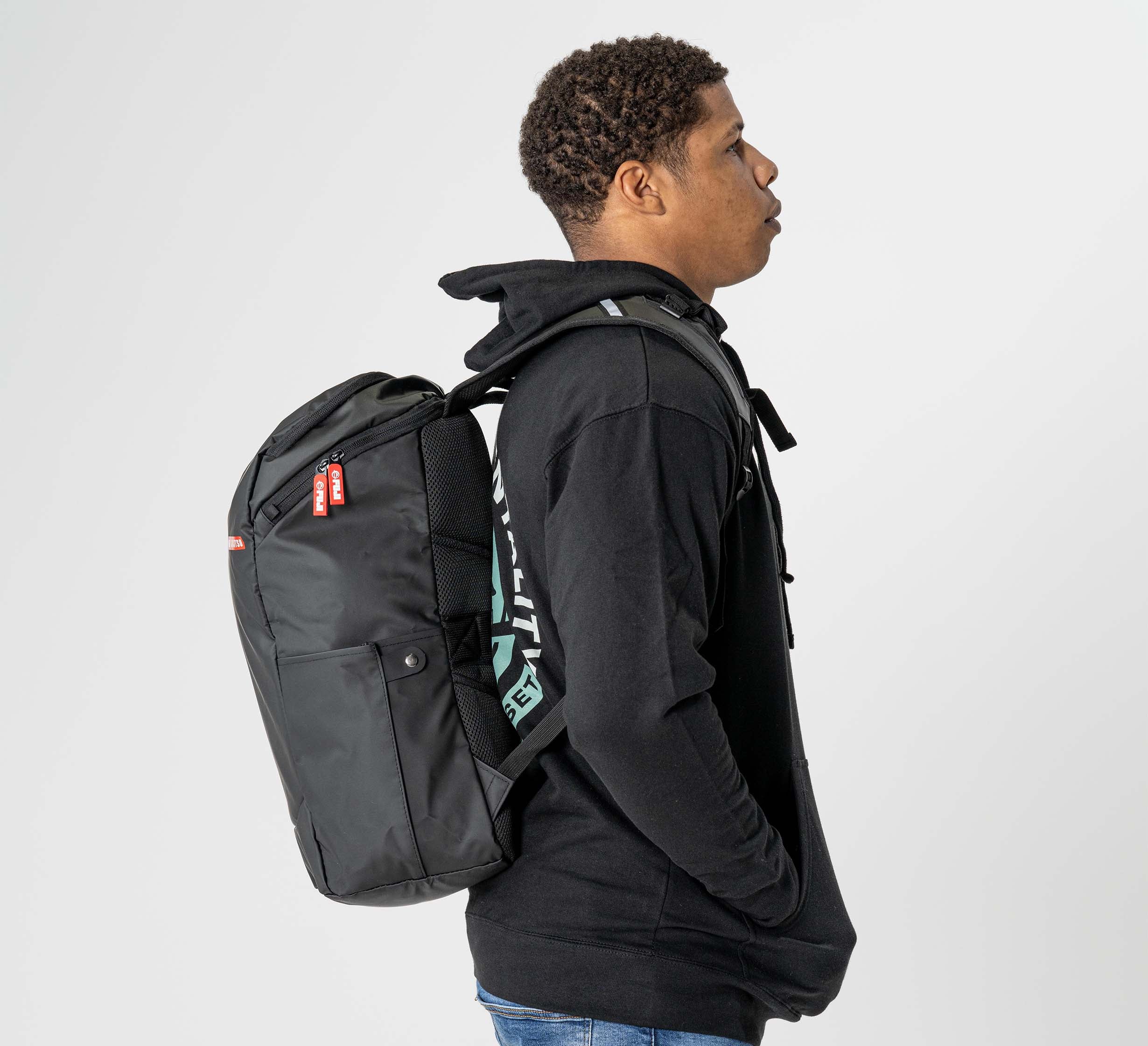 A person in a black hoodie and blue jeans stands in profile against a white background, carrying the Urban Day Backpack Black.