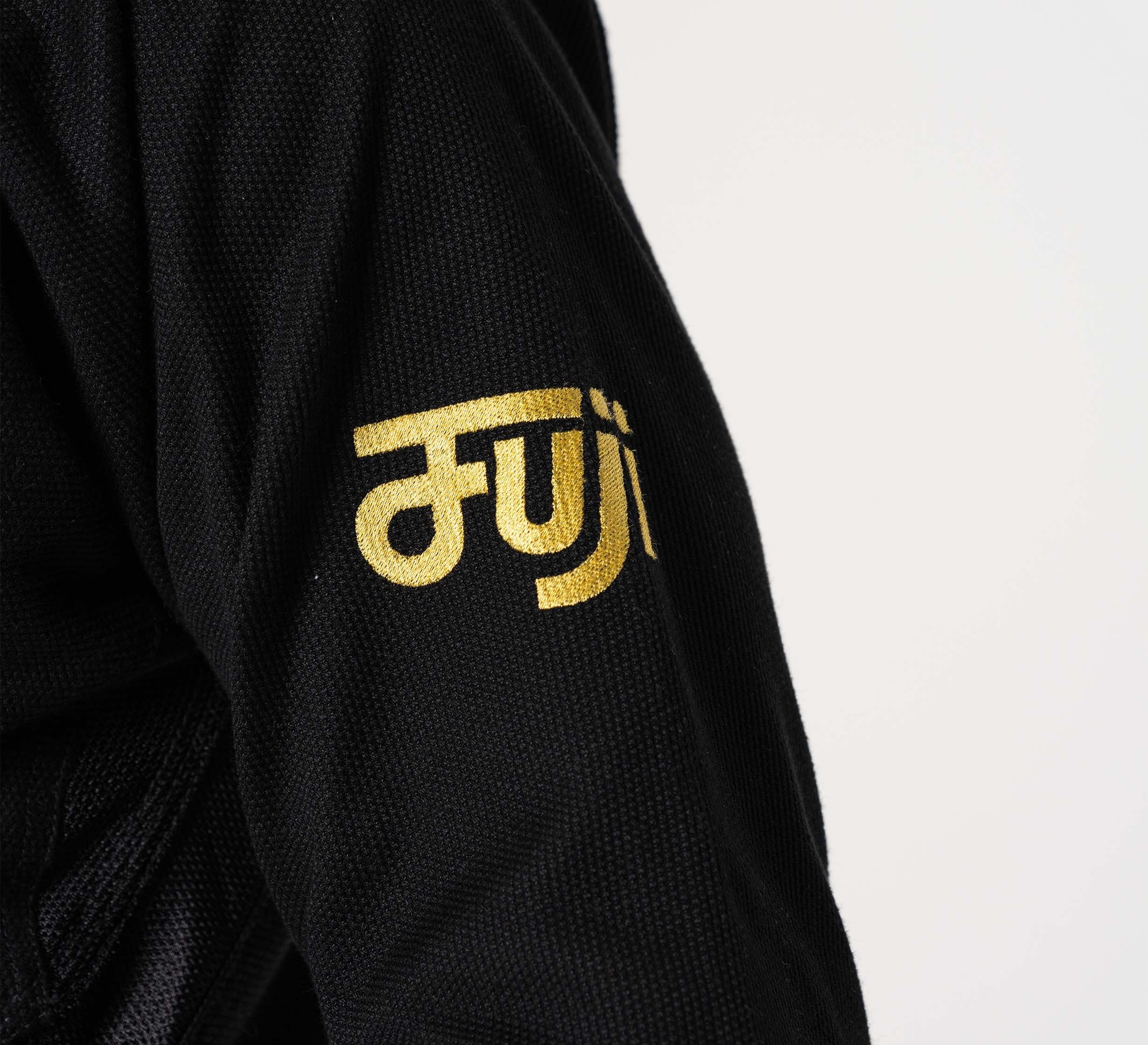 Close-up of a person wearing the Flow-Tech BJJ Gi Black/Gold, featuring bold gold "FUJI" lettering on the sleeve. The background is plain and light-colored.
