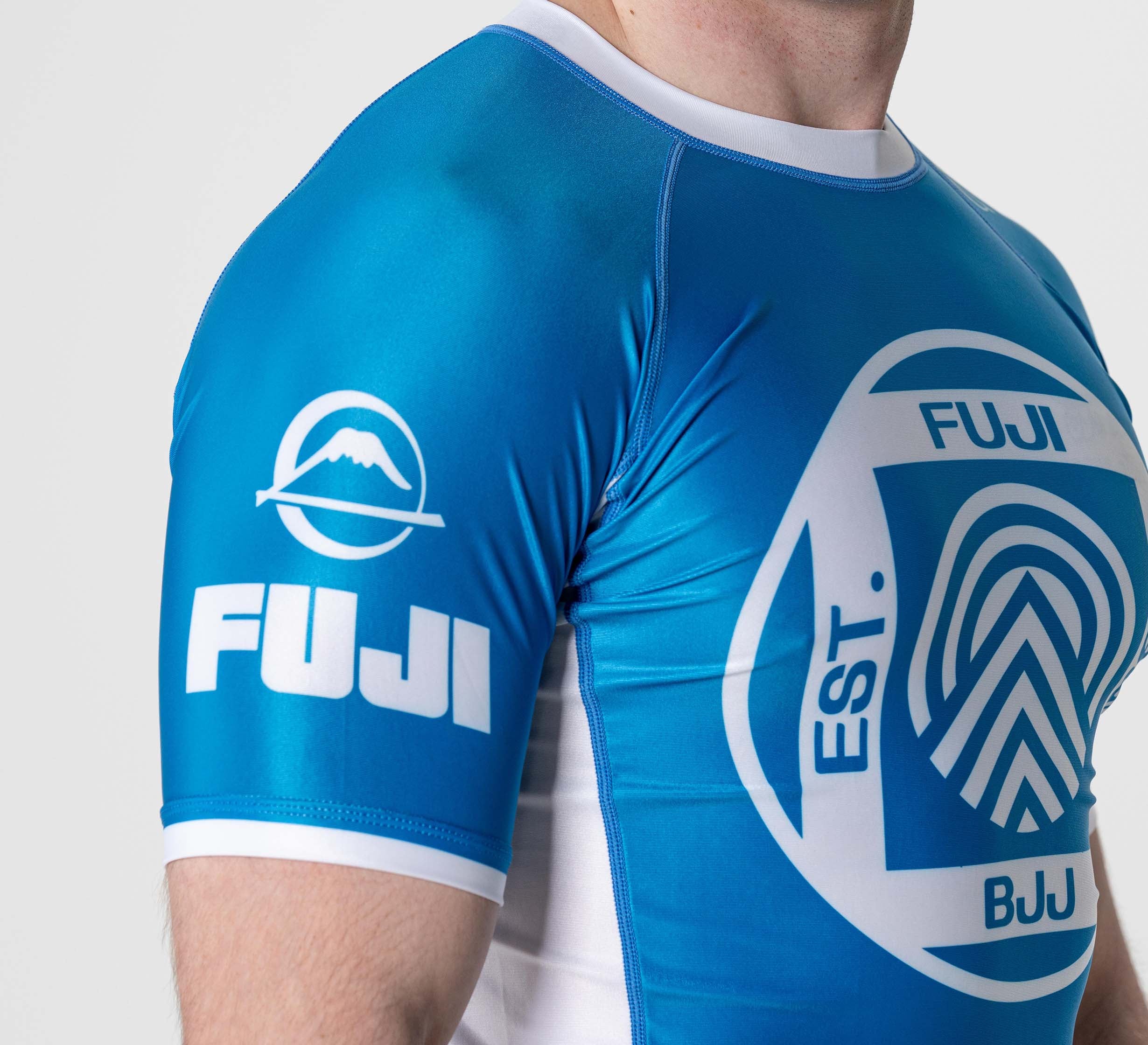 A person wearing the BJJ Flex Lite Rashguard Blue, featuring the FUJI logo and mountain symbol on the sleeve and a large round logo on the chest. The image highlights their upper torso and arm.