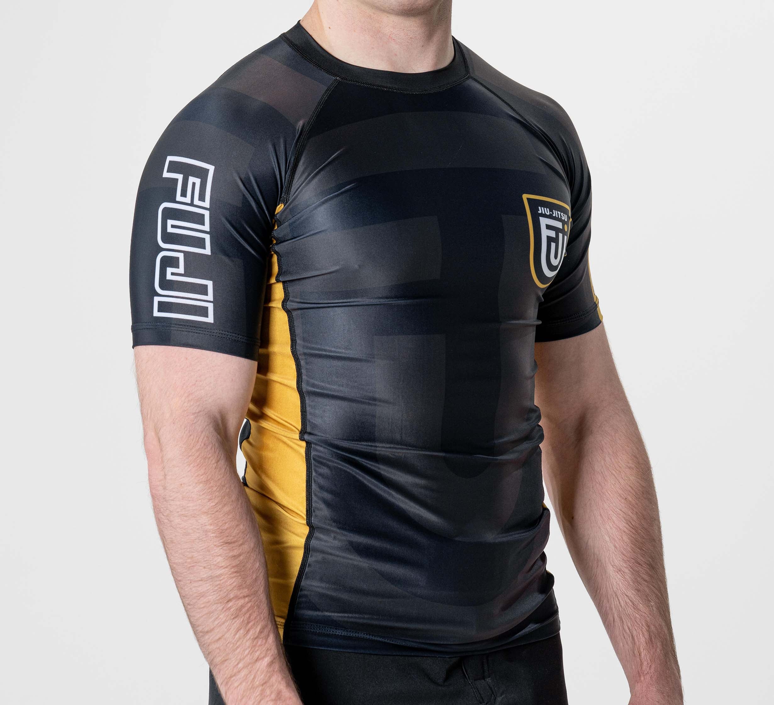 A person is wearing the Rebel Flex Lite Rashguard Black, a fitted short-sleeve athletic top in black, yellow, and gray with a left chest logo and “FUJI” printed in white on the right sleeve. The face is not visible.