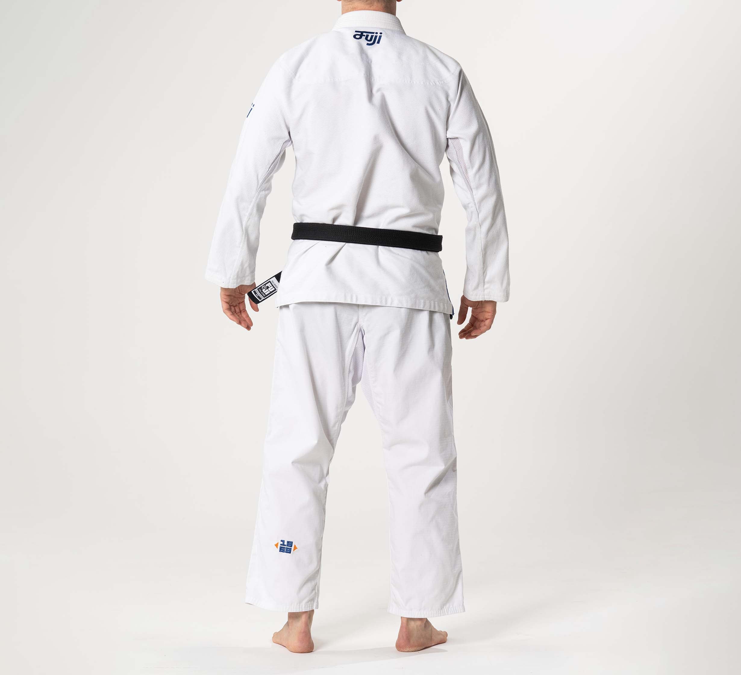 A person stands barefoot, facing away, wearing the Flow-Tech BJJ Gi White/Navy/Orange with a black belt. The back of the gi shows small logos on both the jacket and pants.