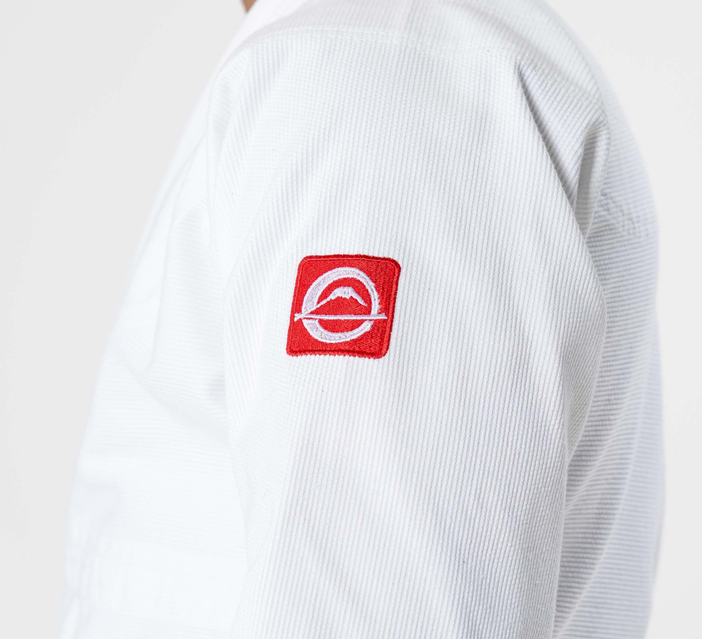 A close-up of a person wearing the Kids Ultra Lite 300 BJJ Gi White, featuring a red patch with a white logo on the upper arm against a plain, light-colored background.