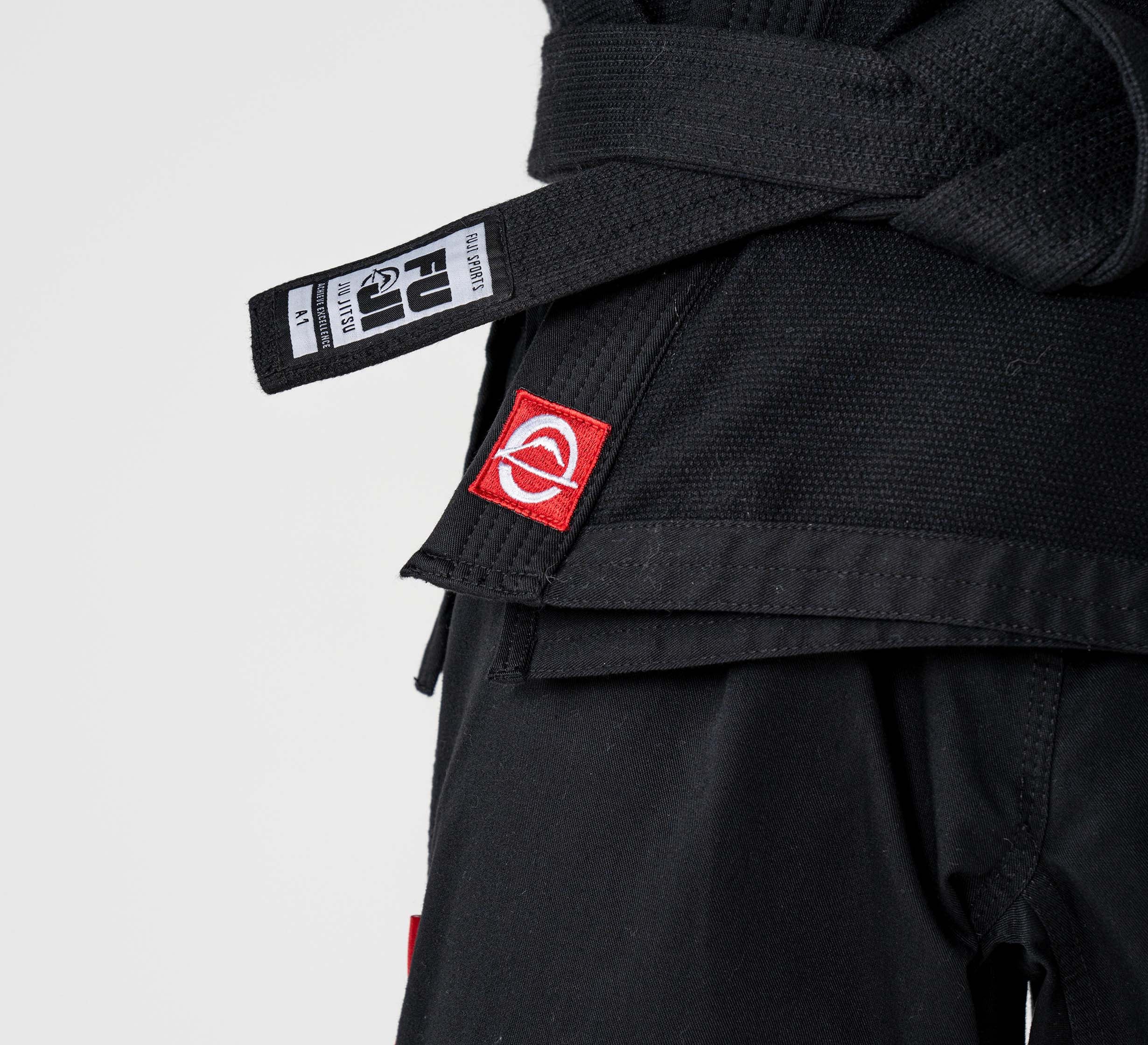 Close-up of a person wearing the Kids Ultra Lite 300 BJJ Gi in black, featuring a white logo on a red patch and FUJI brand tag. The light-colored background highlights the gi’s martial arts style.