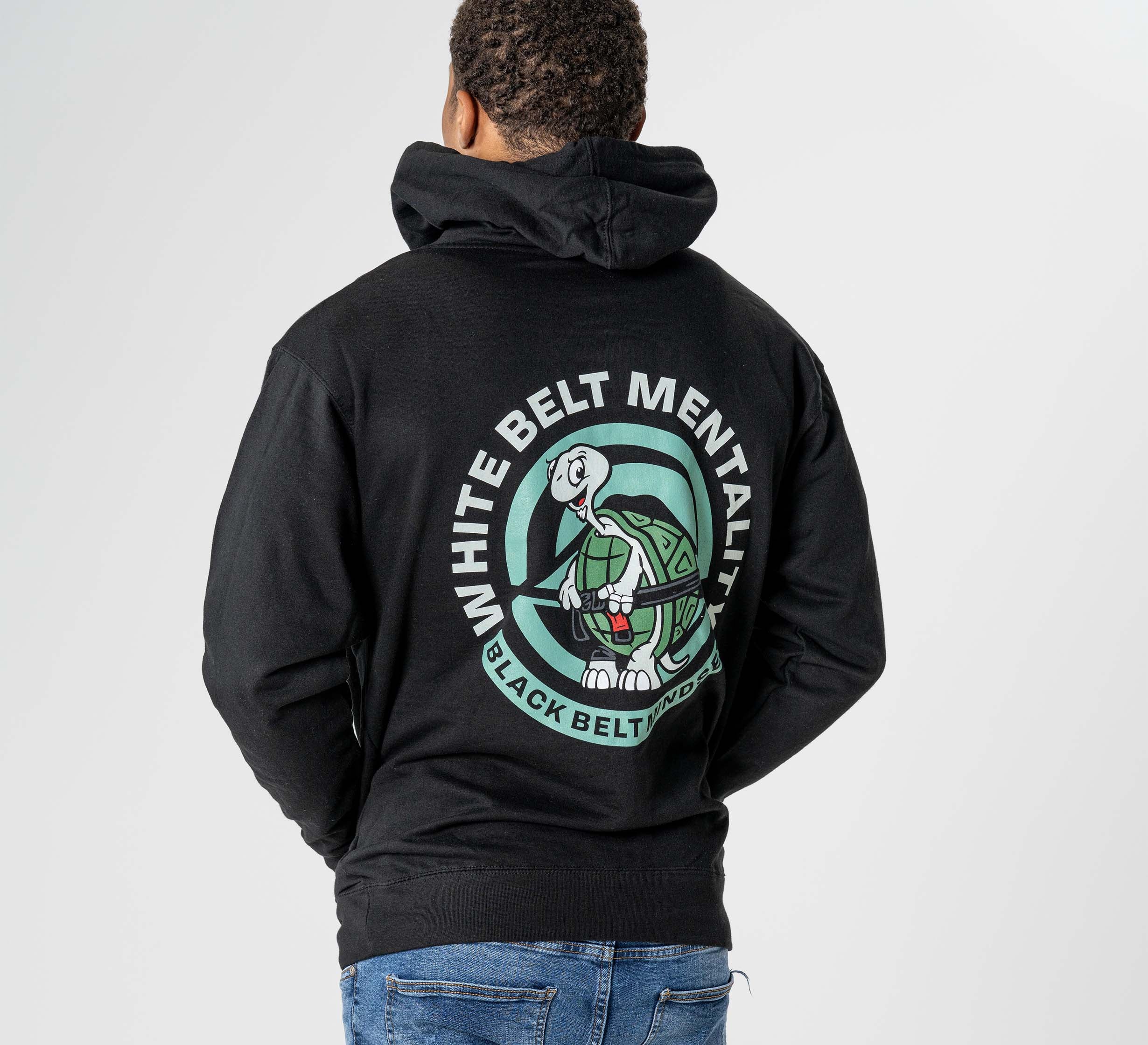 A person is seen from behind wearing the Black Belt Mentality Hoodie Black, featuring a cartoon turtle and the phrases "WHITE BELT MENTALITY" and "BLACK BELT GRIND" in a circular back print.