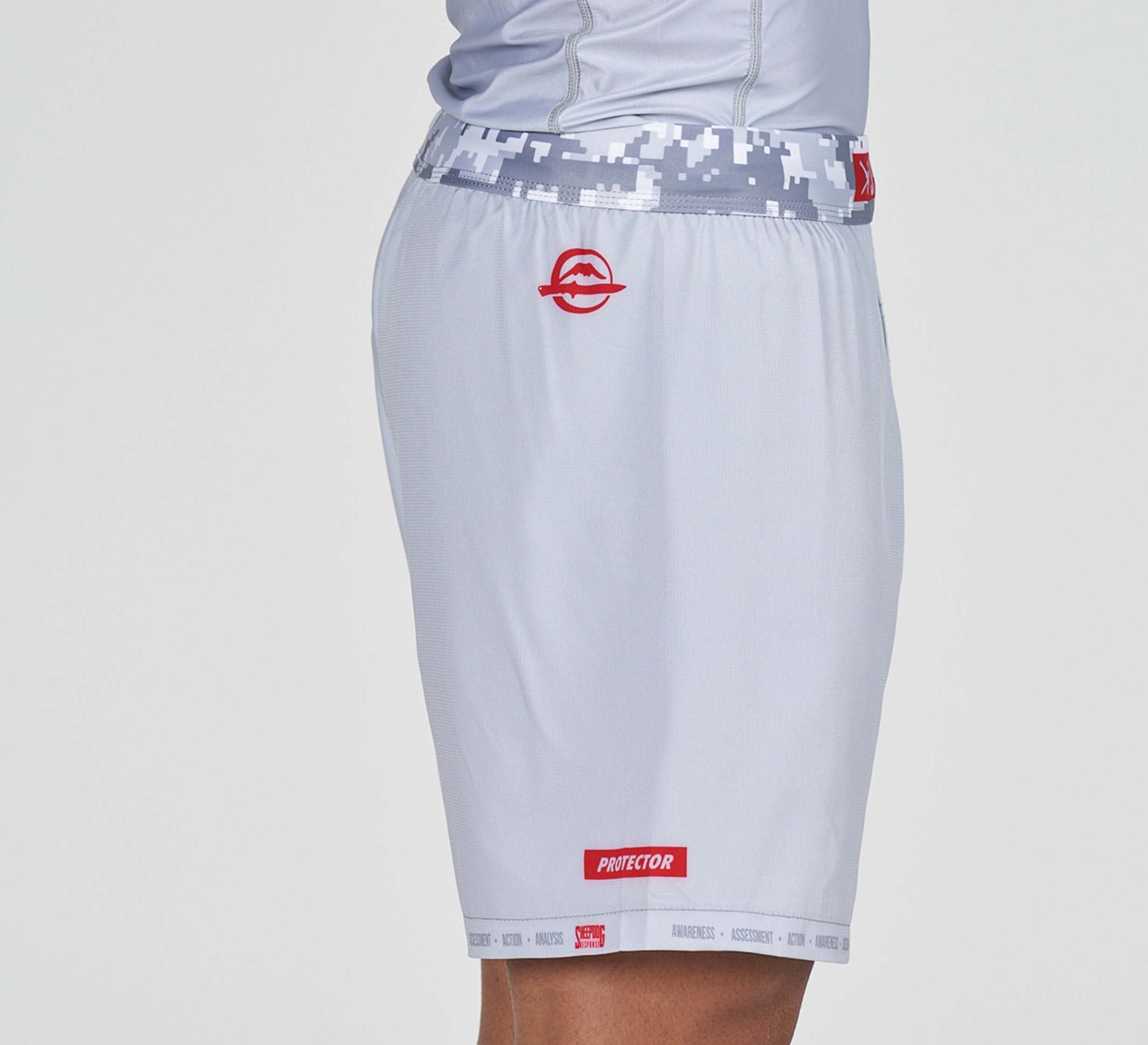 A person wearing Sheepdog Response Flex Lite Shorts Grey, featuring a pixelated gray waistband, red circular logo on the left hip, and “PROTECTOR” in red near the hem. Only the lower torso and upper thigh are visible.