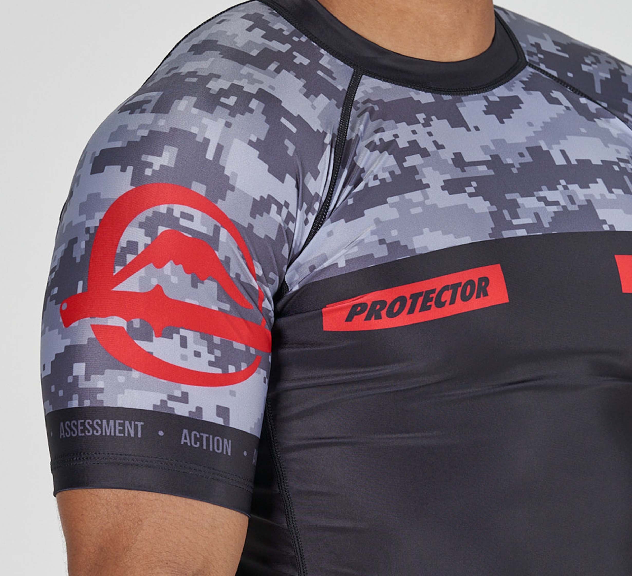 A close-up of a person wearing the Sheepdog Response Flex Lite Rashguard Black, featuring a digital camo pattern, red "PROTECTOR" text, a red sleeve logo, and the words "ASSESSMENT" and "ACTION" on the sleeve.