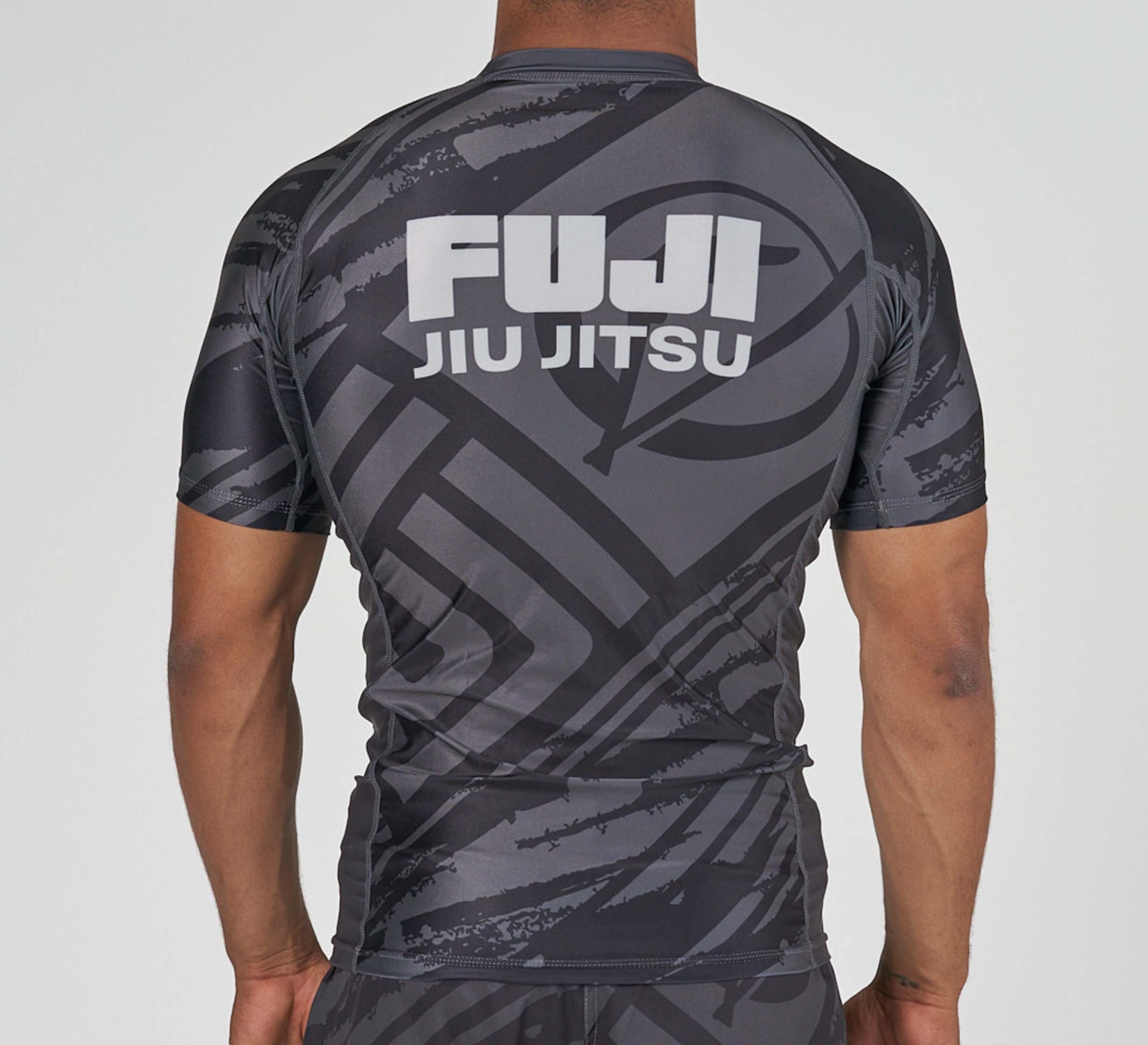 A person wearing the Battle Flex Lite Rashguard Black, featuring short sleeves and "FUJI JIU JITSU" in bold white letters on the back, is shown facing away from the camera.
