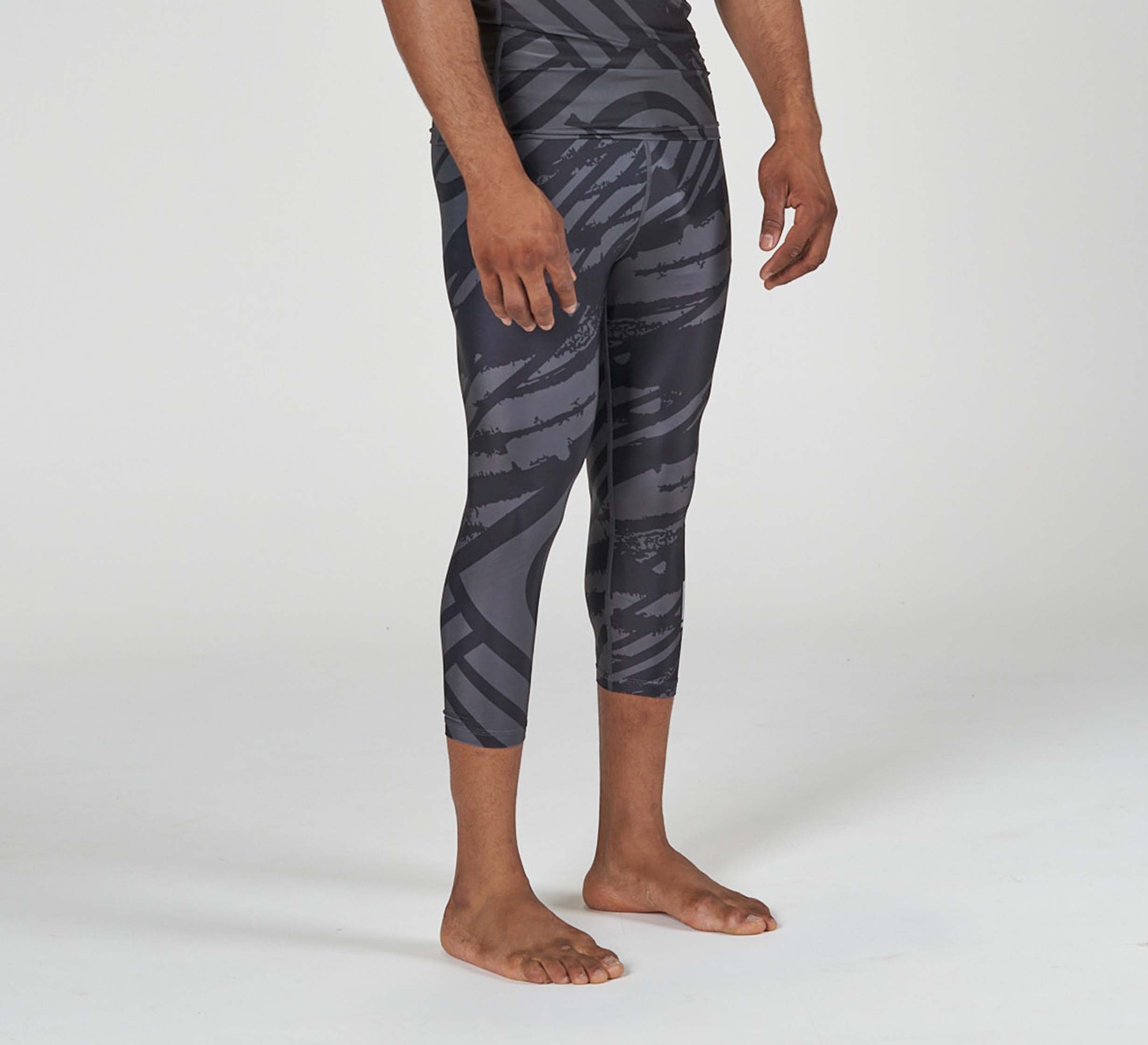 A person stands barefoot wearing Battle Flex Lite Spat Black athletic leggings with a black and grey pattern, extending to mid-calf. The upper body is partially out of frame against a plain light background.
