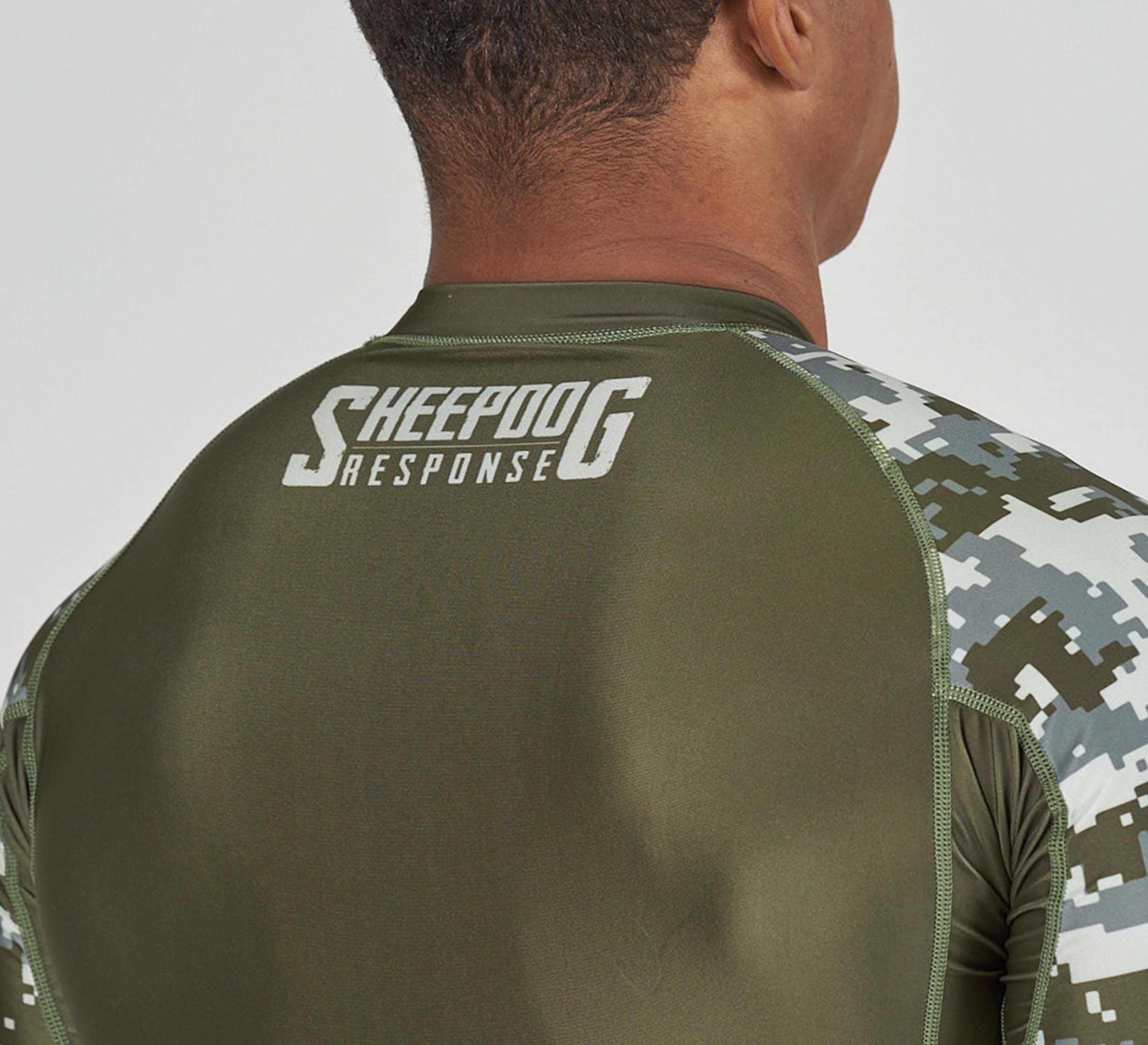 A person wears the Sheepdog Response Flex Lite Rashguard Military Green, featuring "SHEEPDOG RESPONSE" in white on the upper back. The image highlights the athletic shirt's upper back and shoulder area.