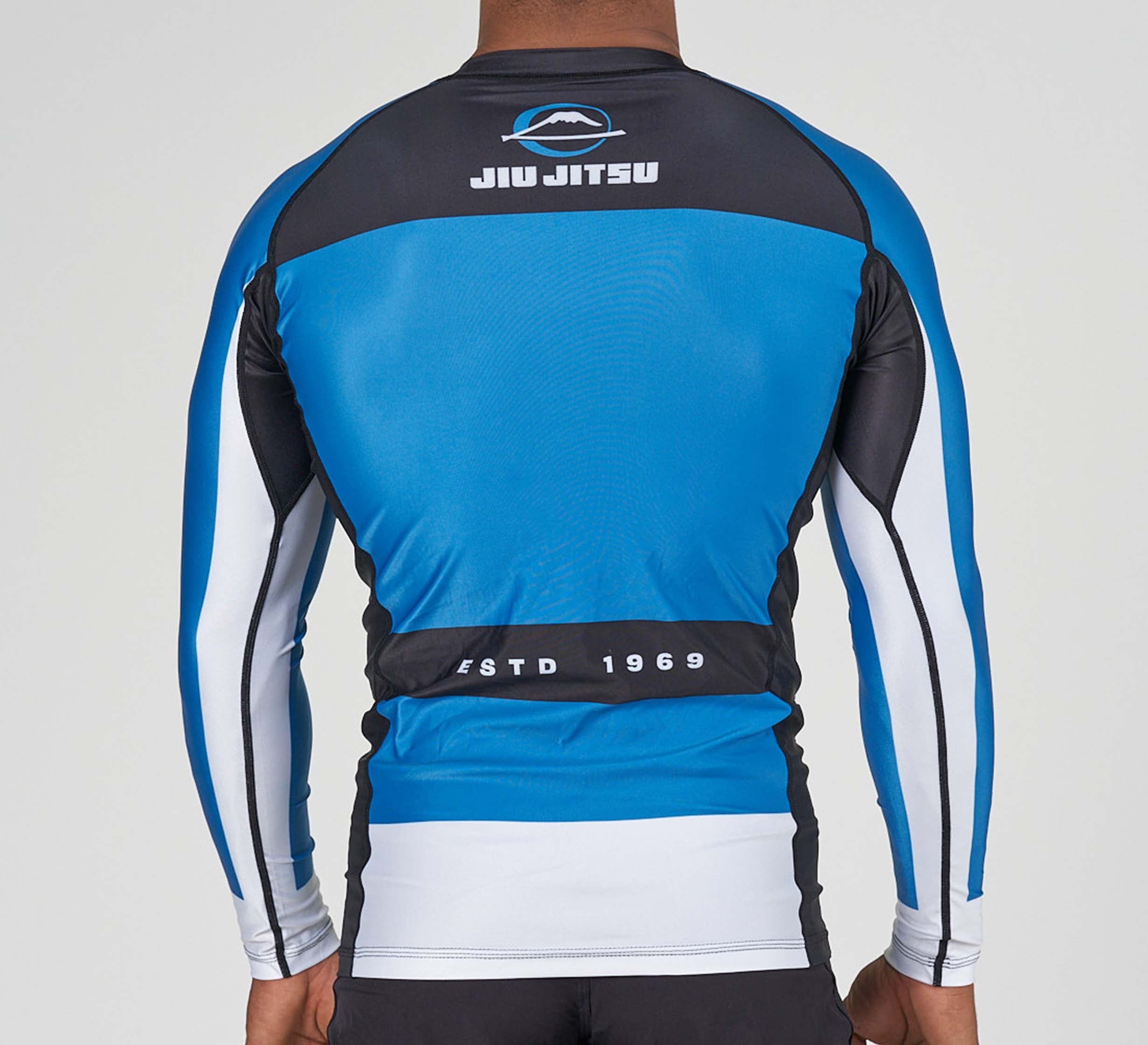 A person is seen from behind wearing the Jiu Jitsu Script Flex Lite Long Sleeve Rashguard Blue, which displays "JIU JITSU" and "ESTD 1969" with a small mountain logo on the back.