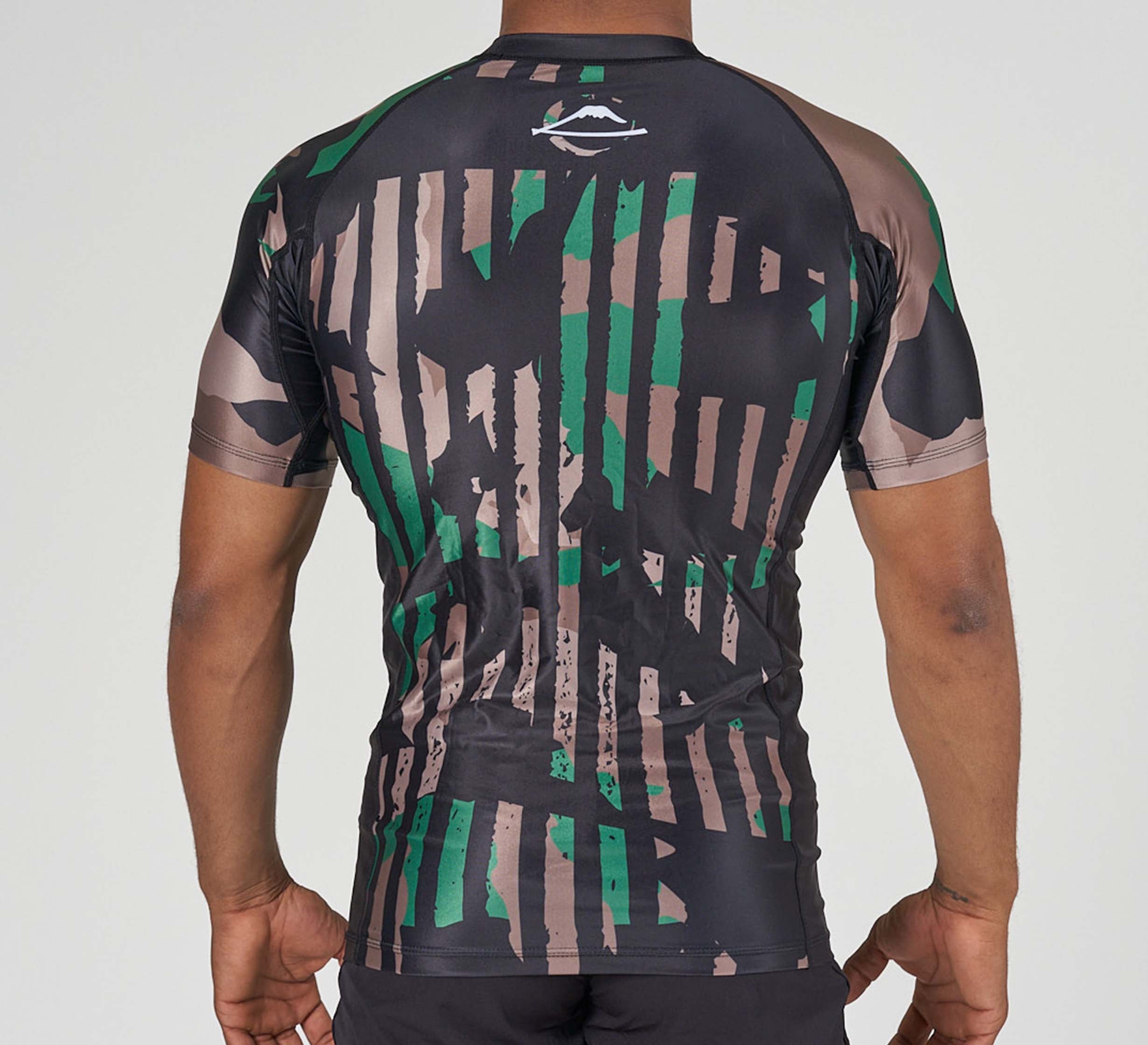 A person seen from the back wears the Camo Flex Lite Rashguard, a fitted short-sleeve athletic shirt featuring an abstract green and beige camo stripe pattern, set against a plain background.